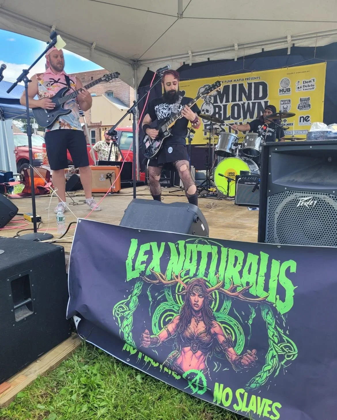 A band performs on an outdoor stage with three members playing guitars and drums. They are under a white canopy and a large yellow banner in the background reads "Mind Blow". A black banner in the foreground displays artwork with the text "Lexiturnalis - No slaves" and features a woman with antlers and mystical symbols.