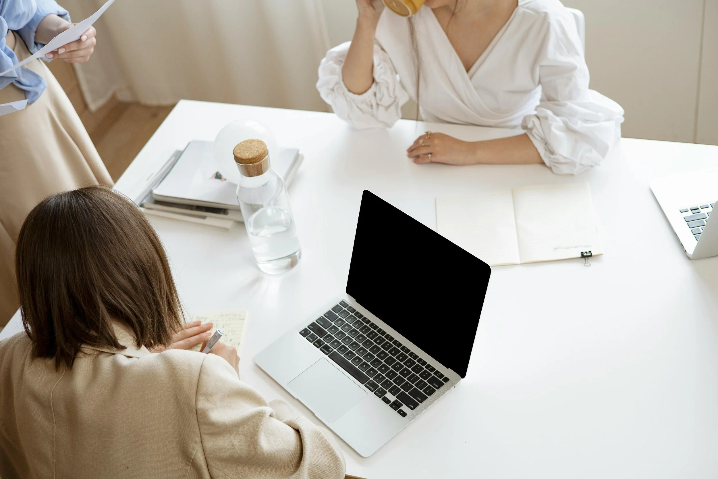 People in a meeting around a white table with laptops, notebooks, a glass of water, and a glass jar. One person is writing in a notebook, another is drinking from a glass, and a third person is holding papers.