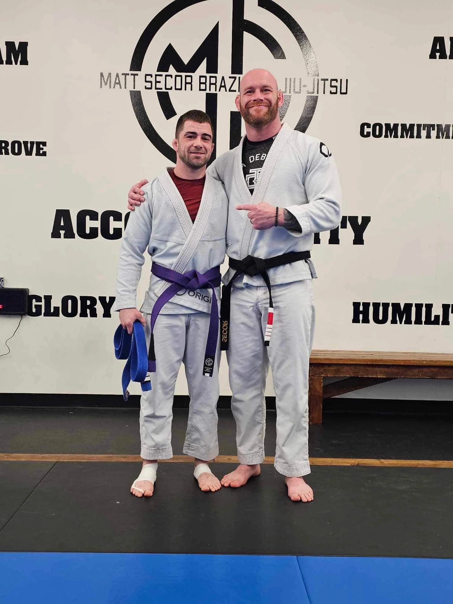 Two men in Brazilian Jiu-Jitsu gis with purple and black belts, standing on a mat in a training facility, smiling with arms around each other, behind a wall with martial arts slogans and a logo.