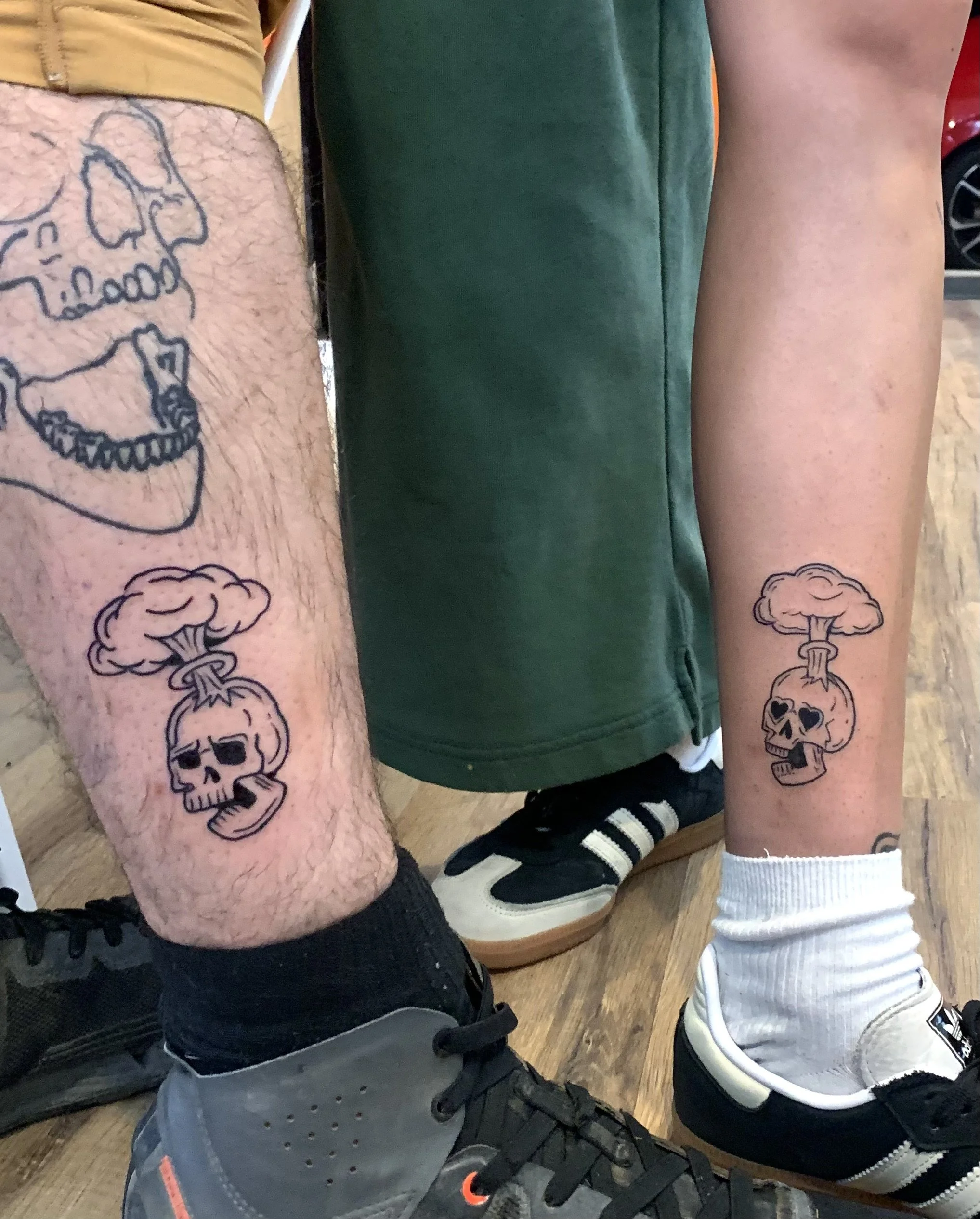 Two individuals with tattoos on their legs, one male and one female, standing side by side on a wooden floor.