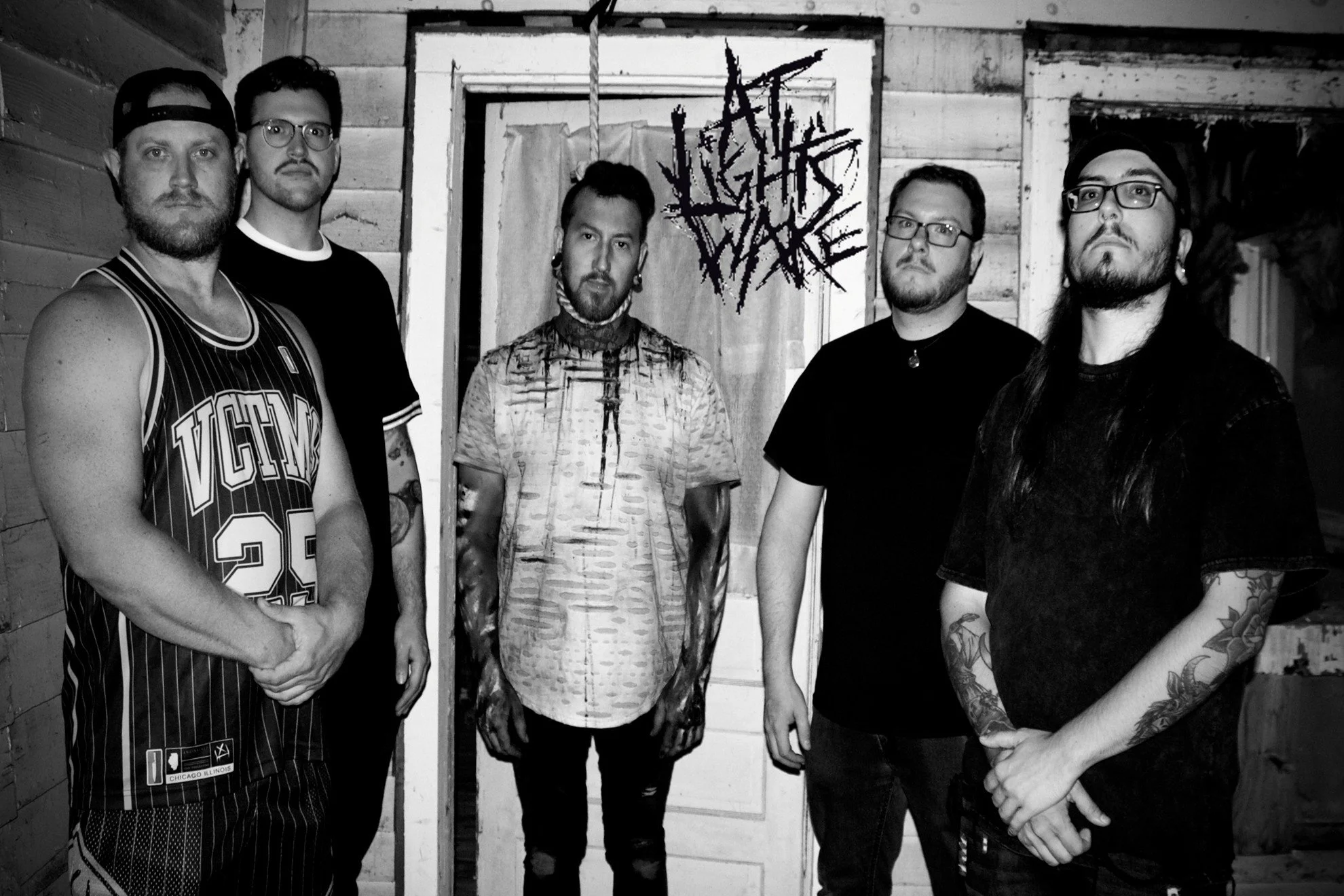 A black and white photo of five men standing in front of a wooden wall and door with a graffiti-style sign that reads 'Death Design' behind them. They have serious expressions and are dressed in casual clothing, with some having tattoos and glasses.