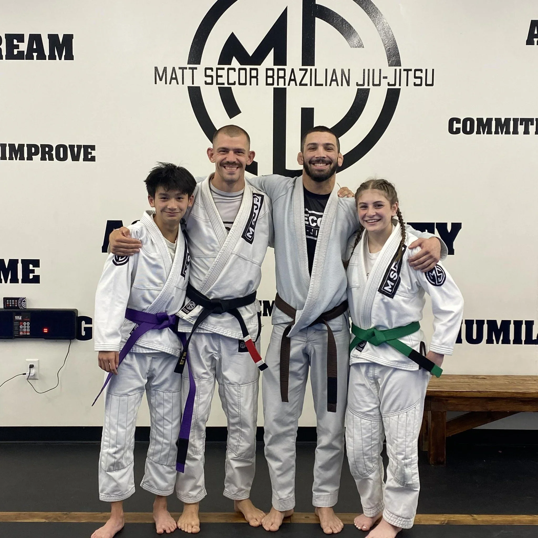 Four people in Brazilian Jiu-Jitsu gis with belts standing together at a martial arts gym. Two young women with purple and green belts, and two men with black and brown belts, smiling with arms around each other. The background features the gym's logo and motivational words.