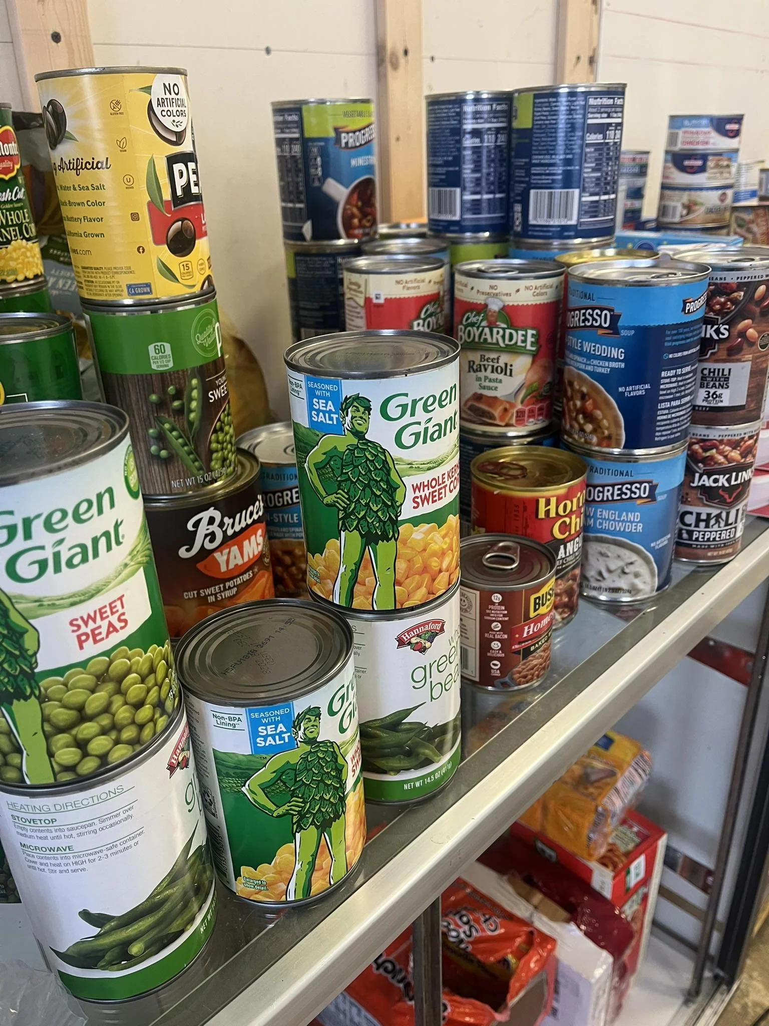 Shelf of canned goods including green giant sweet peas, sea salt green beans, Bush's baked beans, Hormel chili, Progresso soup, and Chef Boyardee ravioli.