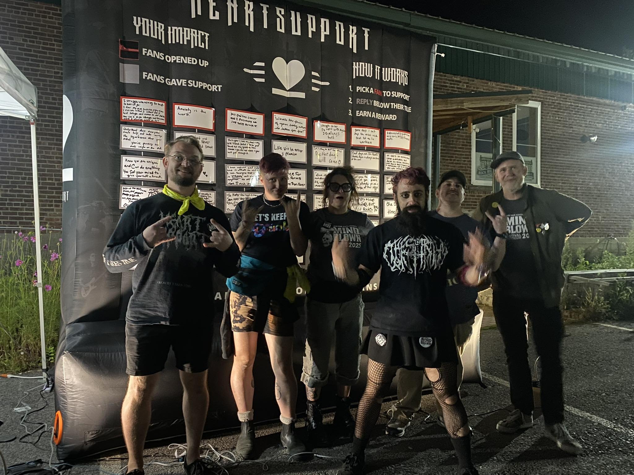 A group of six people posing in front of a large support board at night. They are making rock hand gestures and smiling. The support board has various messages and information, including a header that reads 'Support' and 'How it works,' with steps for supporting fans. The area is outdoors with some plants and a brick wall visible.