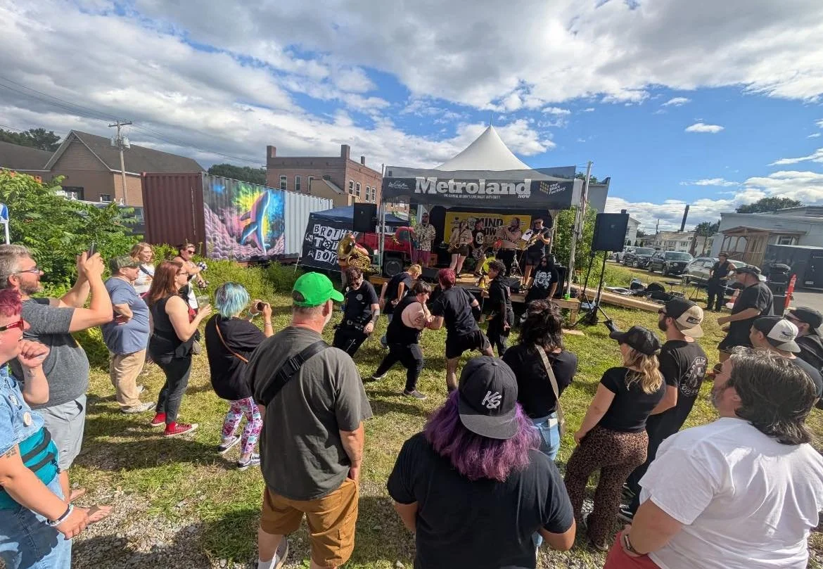 People enjoying a live outdoor concert, dancing and taking photos, with a band playing on a stage under a white canopy, on a partly cloudy day.