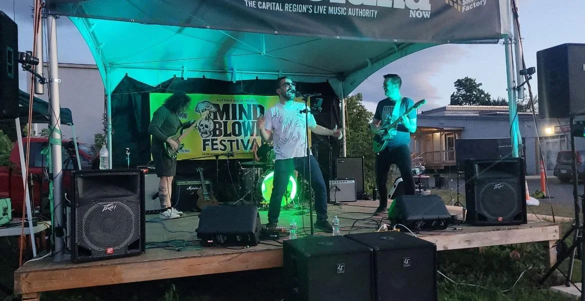 A band performs on an outdoor stage at the Mind Blow Festival during the evening. The band includes a singer, a guitarist, a bassist, and a drummer. The stage is covered by a canopy and is illuminated with blue and green lighting. The background has a band banner and there are speakers, microphones, and water bottles on stage.