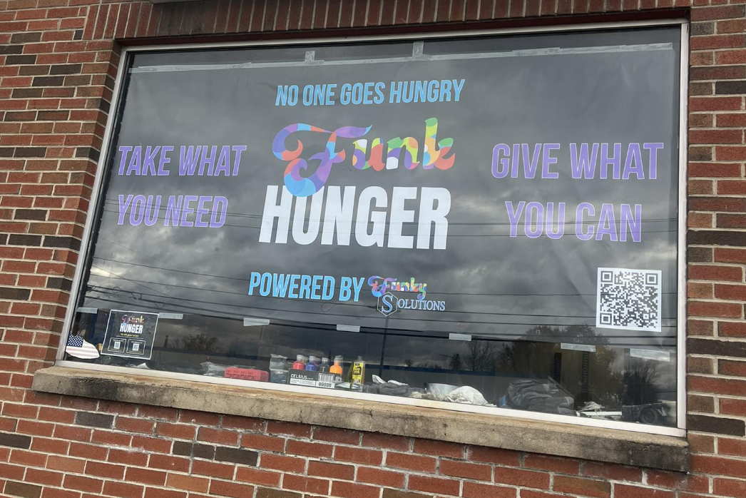 A window display with a colorful sign promoting a food bank or community service. The sign reads: 'NO ONE GOES HUNGRY' at the top, 'THANK YOU' in large, multicolored letters in the center, and 'HUNGER' below it. It also has the phrases 'TAKE WHAT YOU NEED' on the left and 'GIVE WHAT YOU CAN' on the right, along with 'POWERED BY' and a logo for 'Funky Solutions'. There is a QR code on the right side of the window, and some items and signs visible on the window ledge.