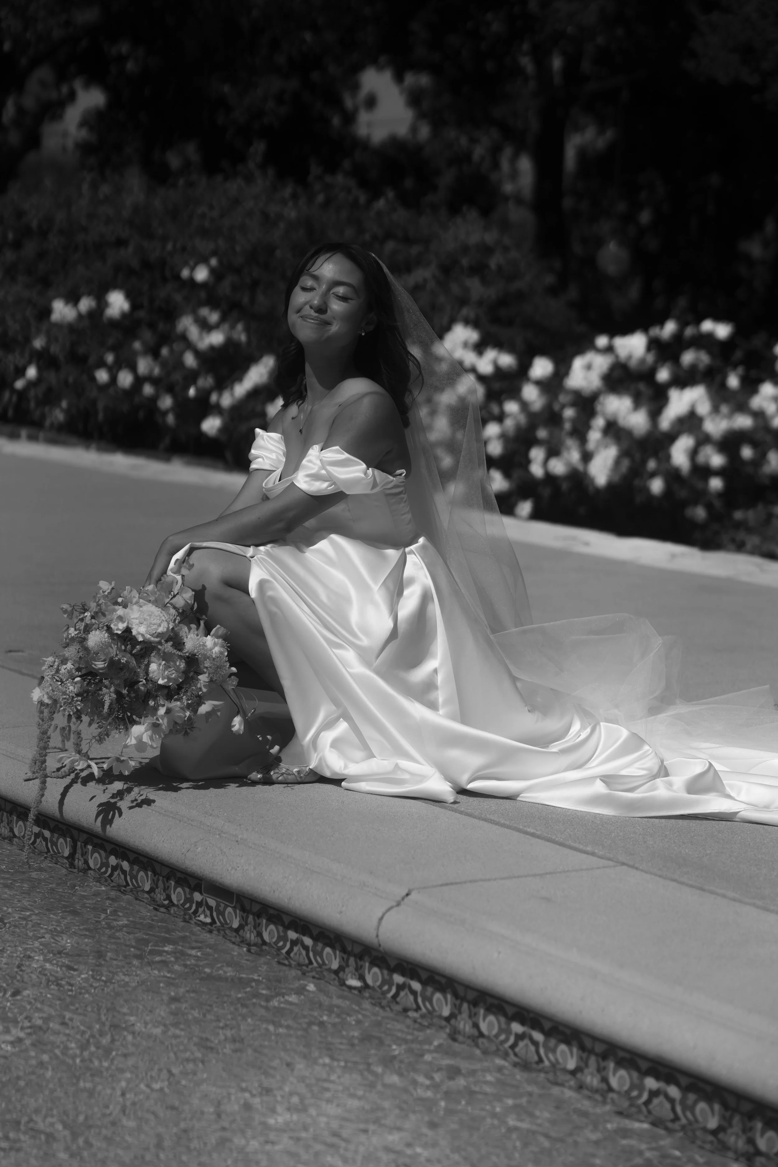 A woman dressed as a bride, sitting on the edge of a pool with a bouquet of flowers, her eyes closed, smiling, wearing a wedding gown and veil.