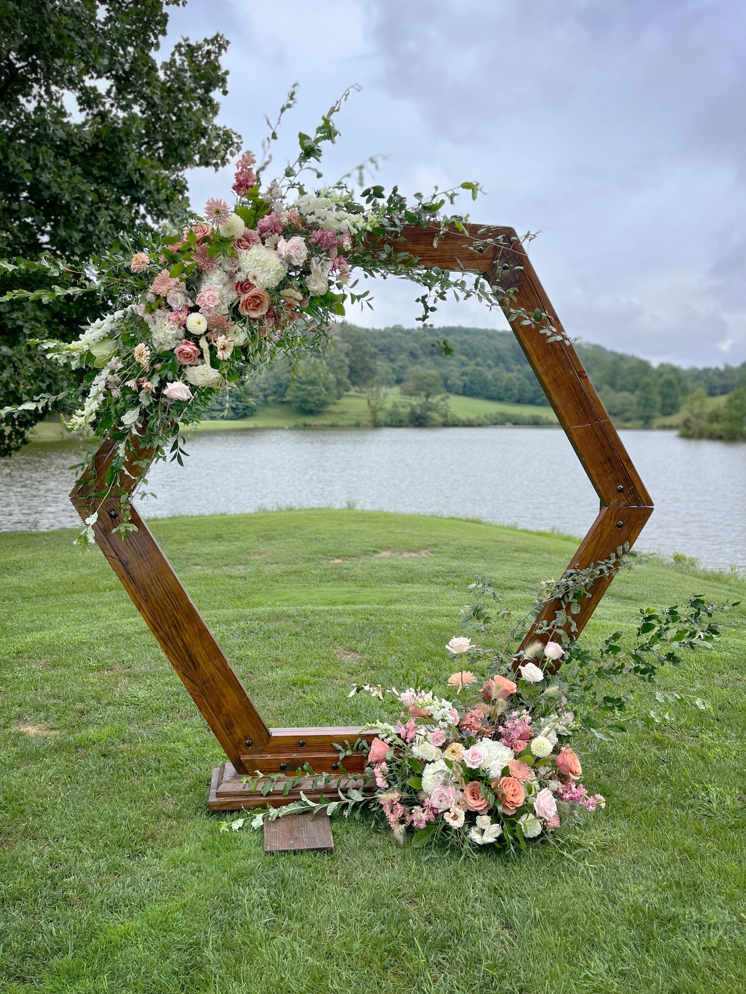 A wooden hexagonal wedding arch decorated with pink, white, and peach flowers and greenery, set on a grassy area by a lake with trees and hills in the background.