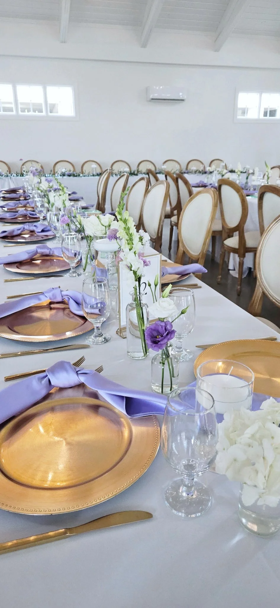 Elegant banquet table with gold chargers, lavender napkins, clear glassware, and white and purple floral centerpieces in a bright, spacious room with white walls and high ceiling.