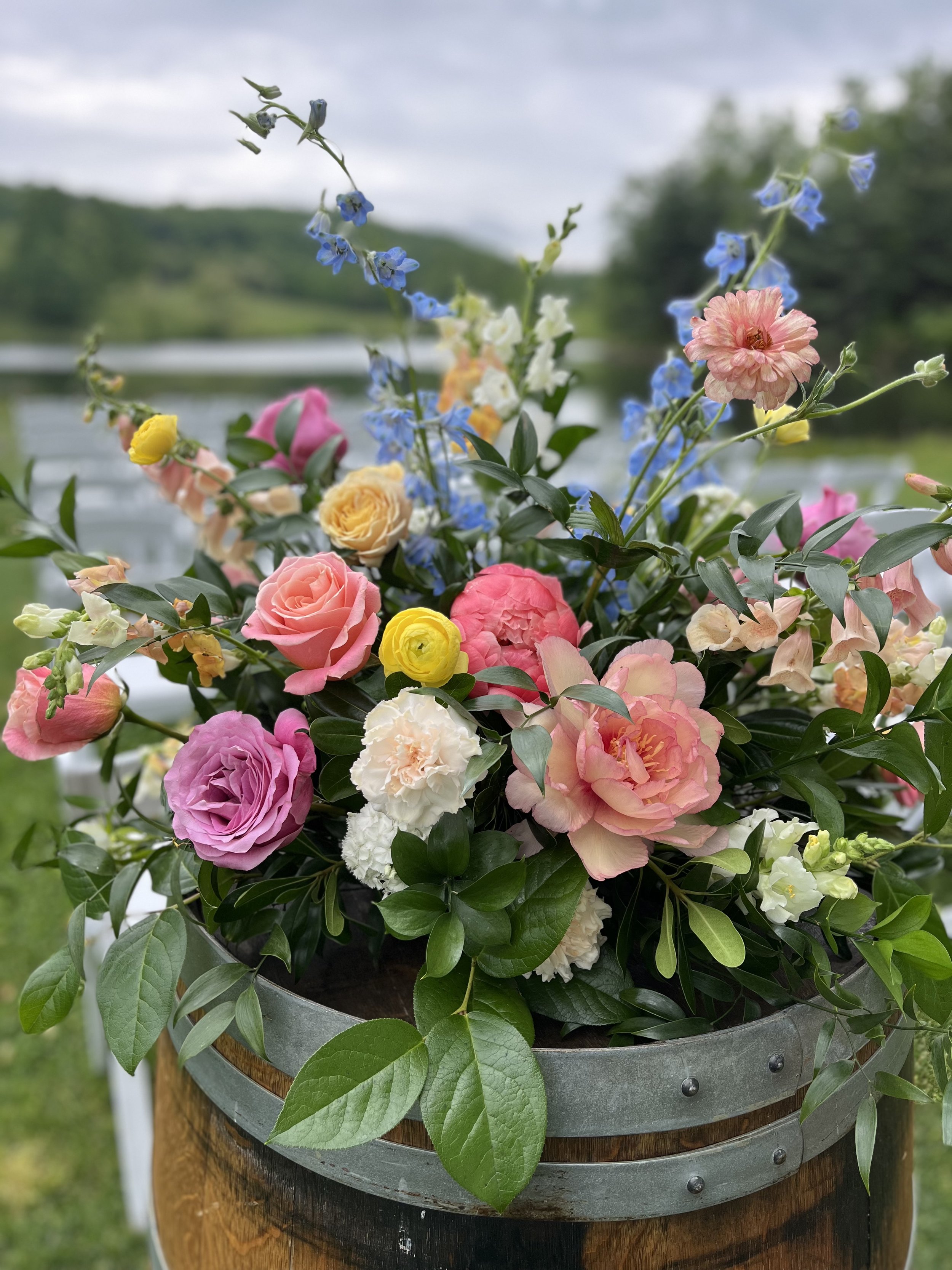 Colorful mixed flower bouquet with roses, carnations, and other flowers in a metal and wood container outdoors near a lake, with a cloudy sky and green landscape in the background.