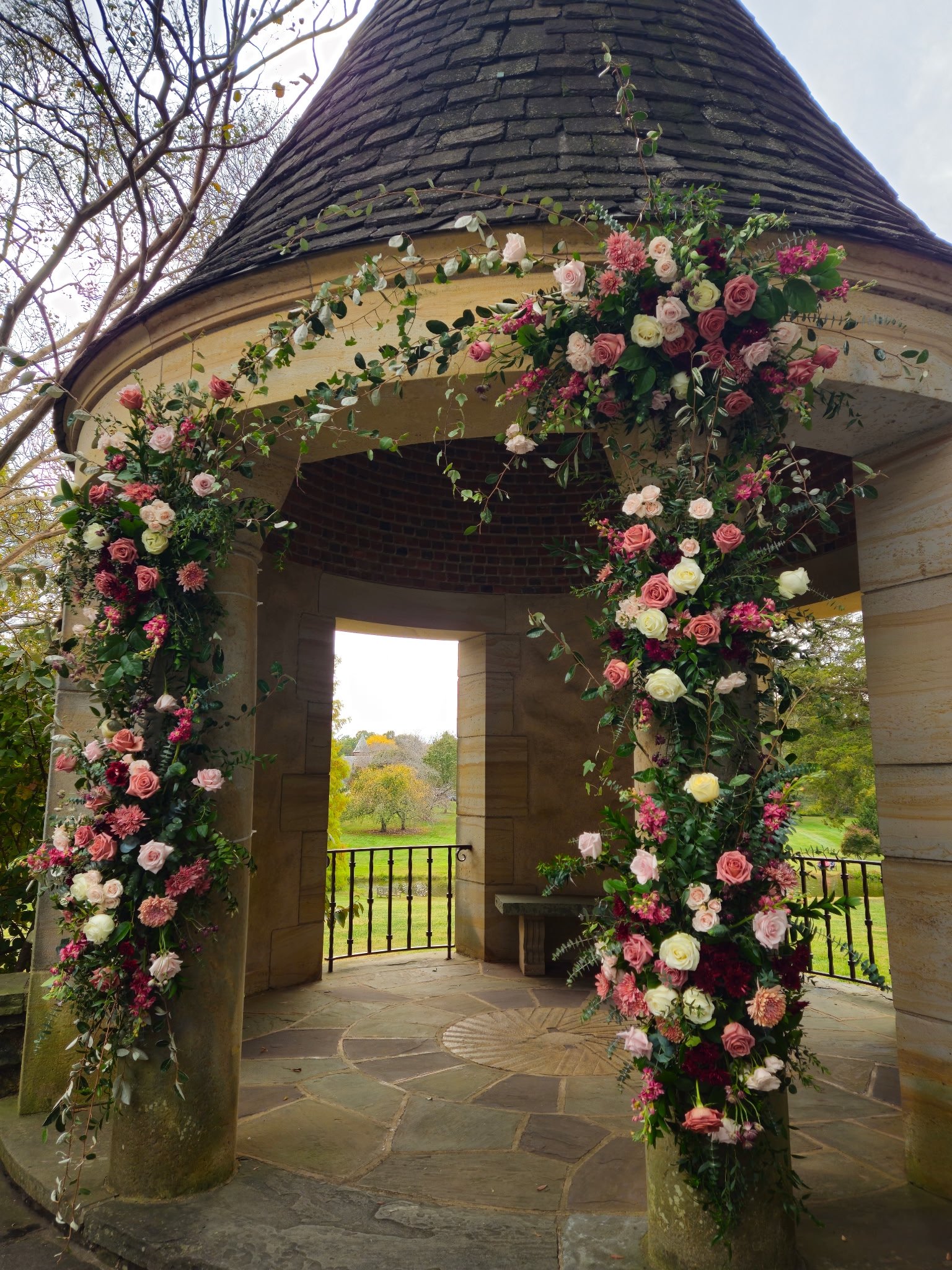A floral archway with pink, white, and purple flowers at a gazebo structure in a park.
