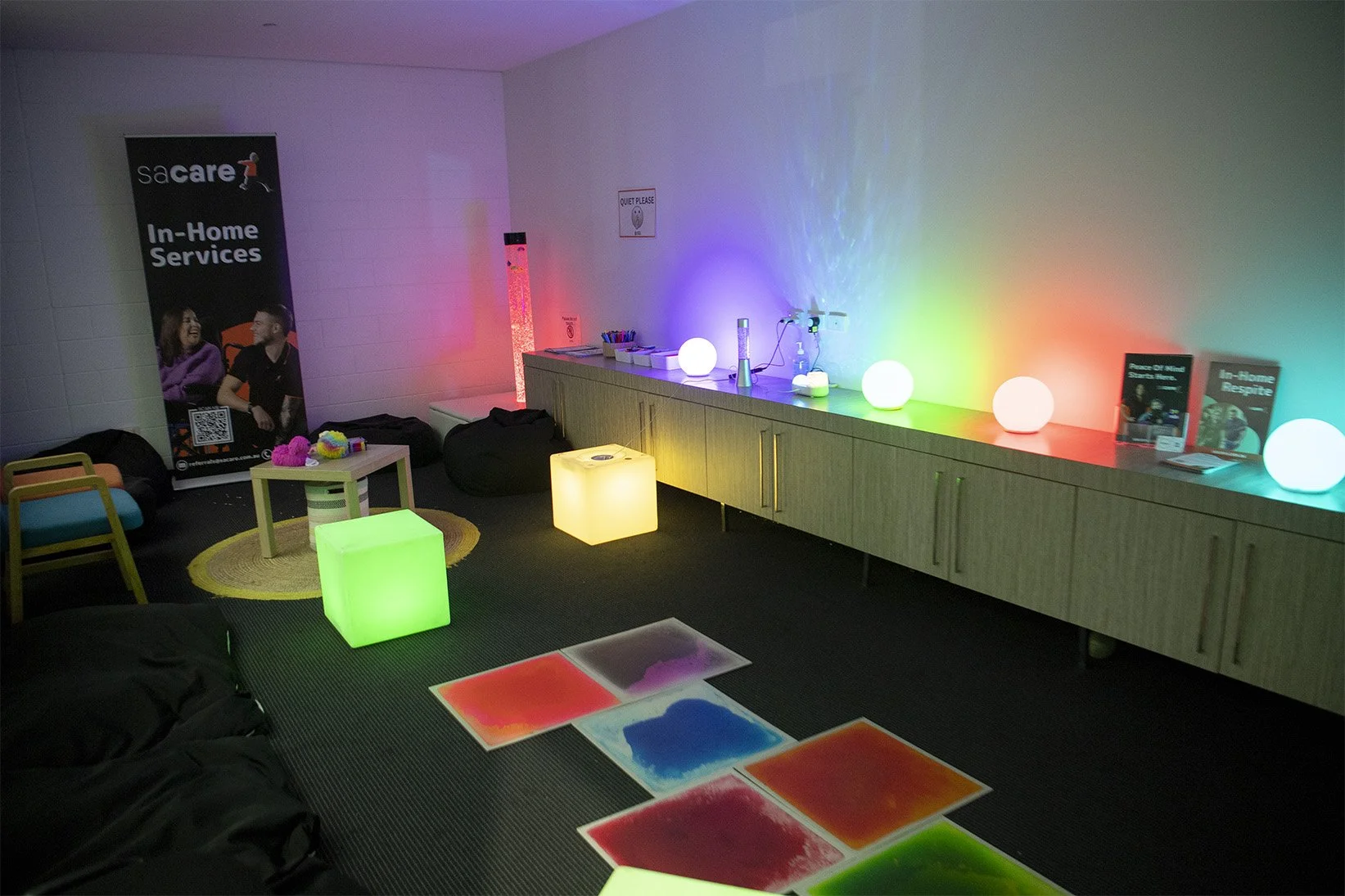 A room with colorful, glowing LED lights on tables, on the floor, and on a counter. There are chairs, bean bags, and art supplies, with a sign on the wall that says "QUIET PLEASE." There is a banner promoting in-home services on one side of the room.