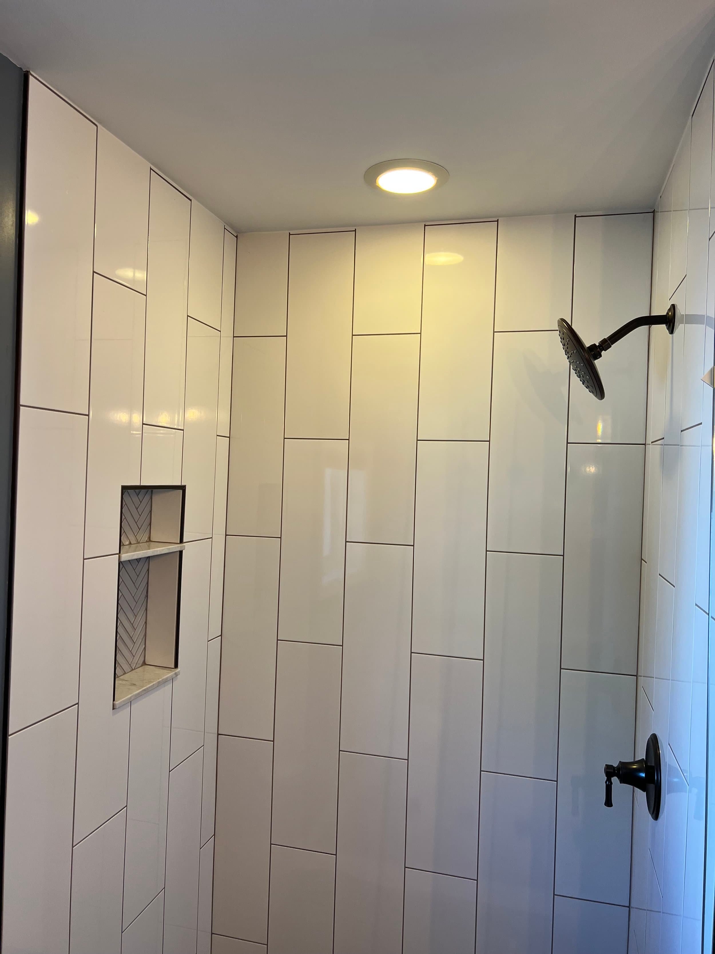 A bathroom shower area with large white rectangular tiles on the walls, a round ceiling light, a black showerhead, a wall-mounted soap holder, and a corner shelf with mosaic tile backing.