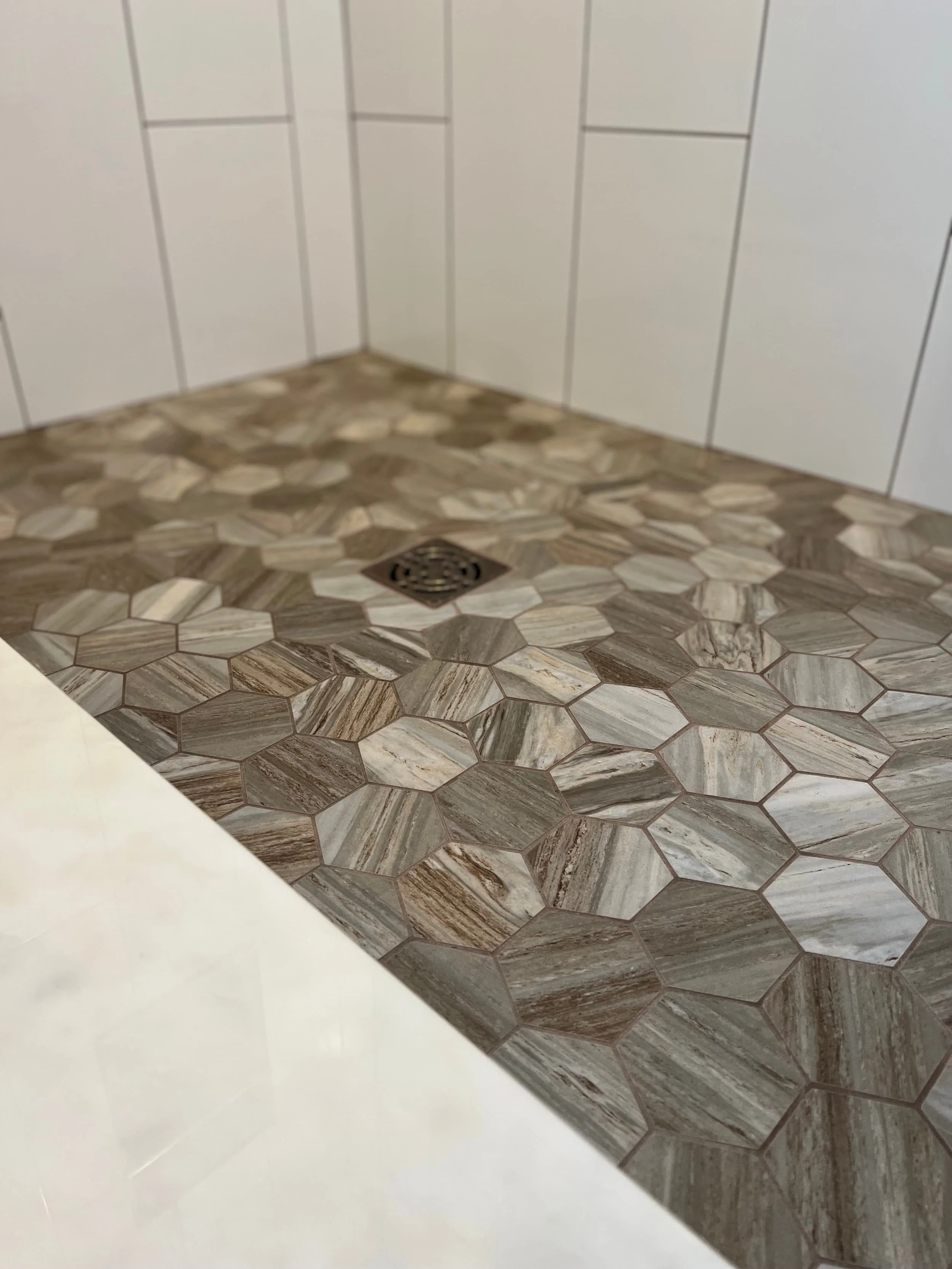 A corner of a room with tiled flooring and white paneled walls. The tiles are hexagonal with a marble pattern in shades of brown, beige, and white. There is a floor drain in the middle of the tiles.