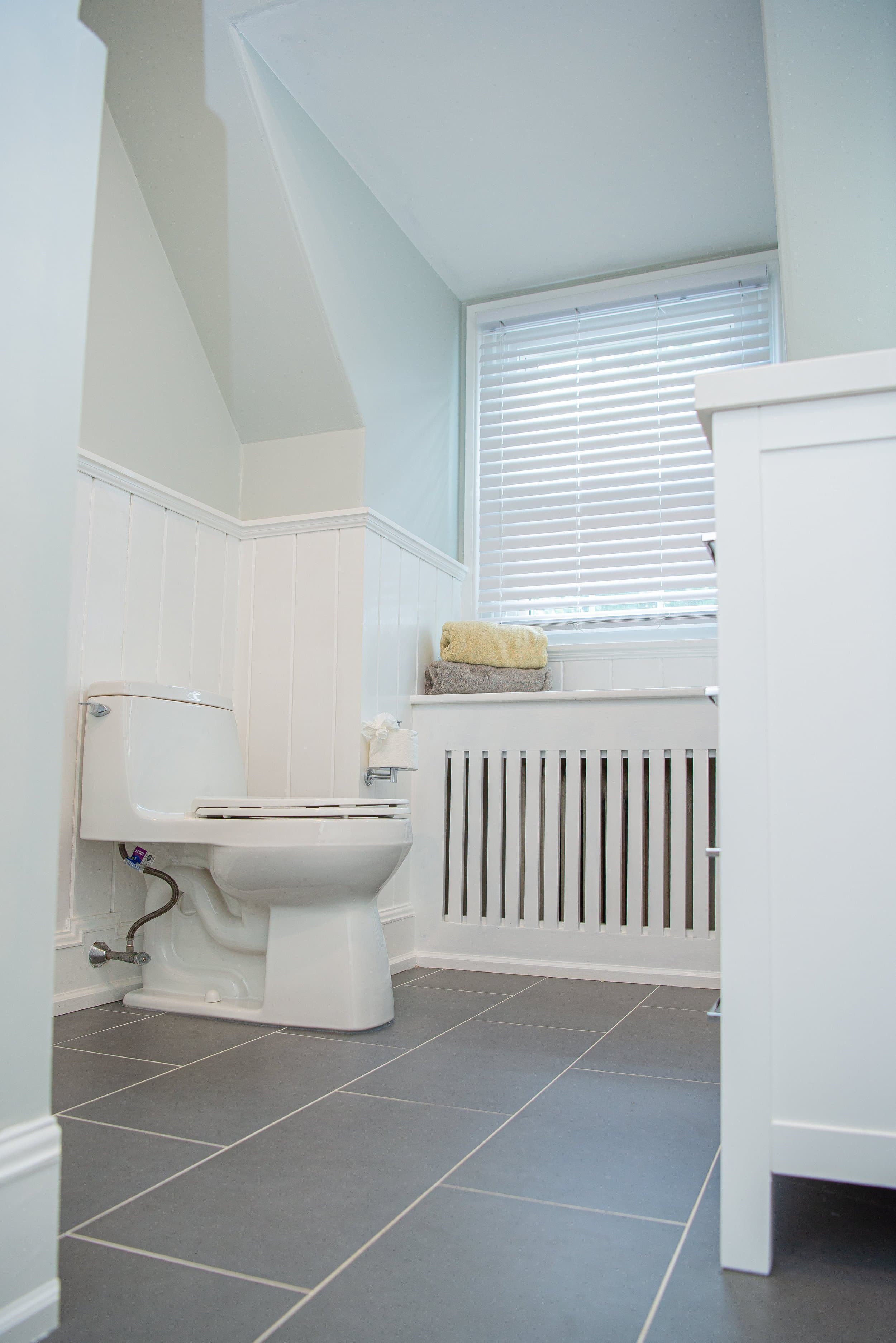 Bathroom with gray tiled floor, white paneled walls, small window with blinds, toilet, and towel on shelf