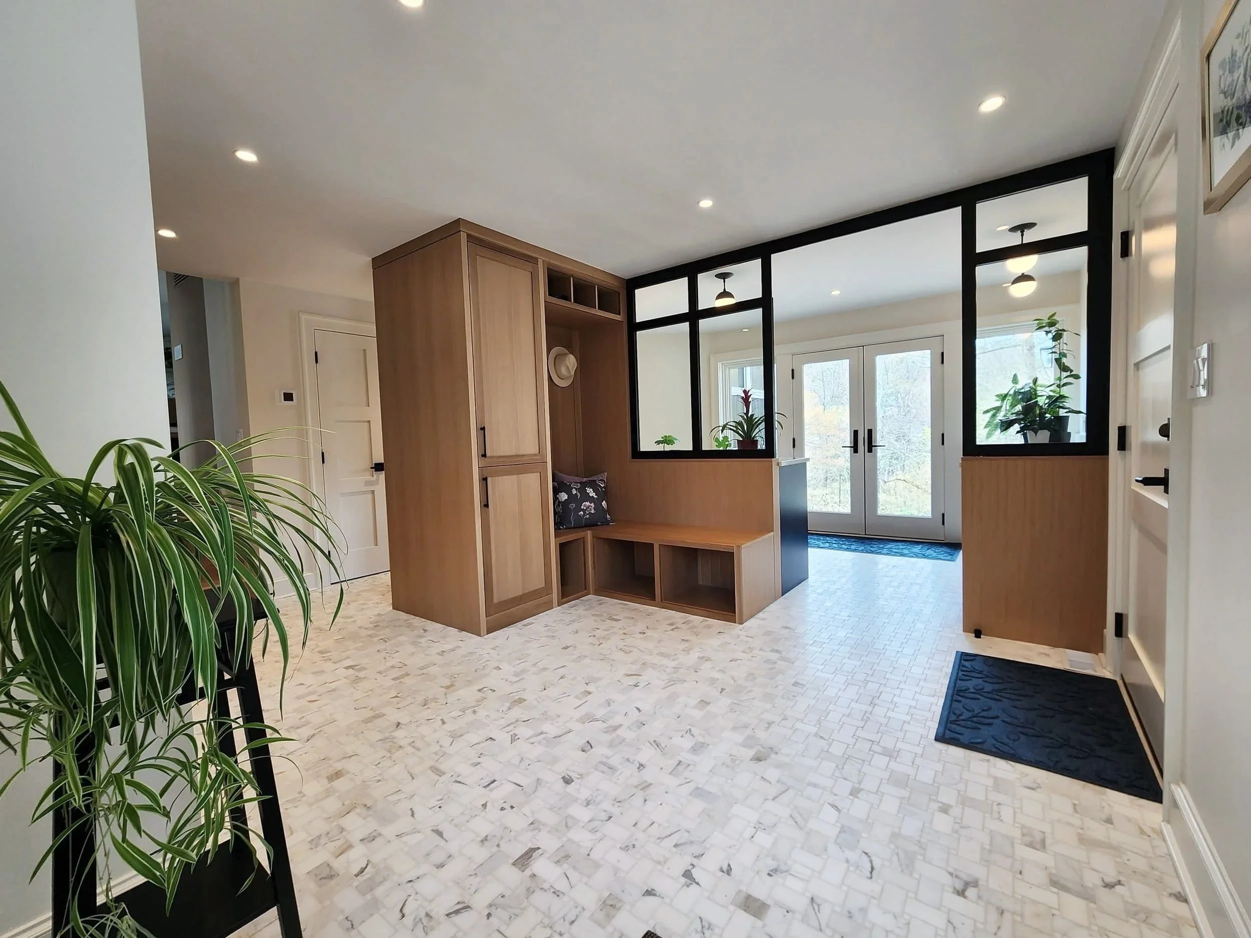 Interior entryway of a house with white and wood accents, black-framed glass doors, potted plants, and a seating nook with a hat on a built-in wooden bench.