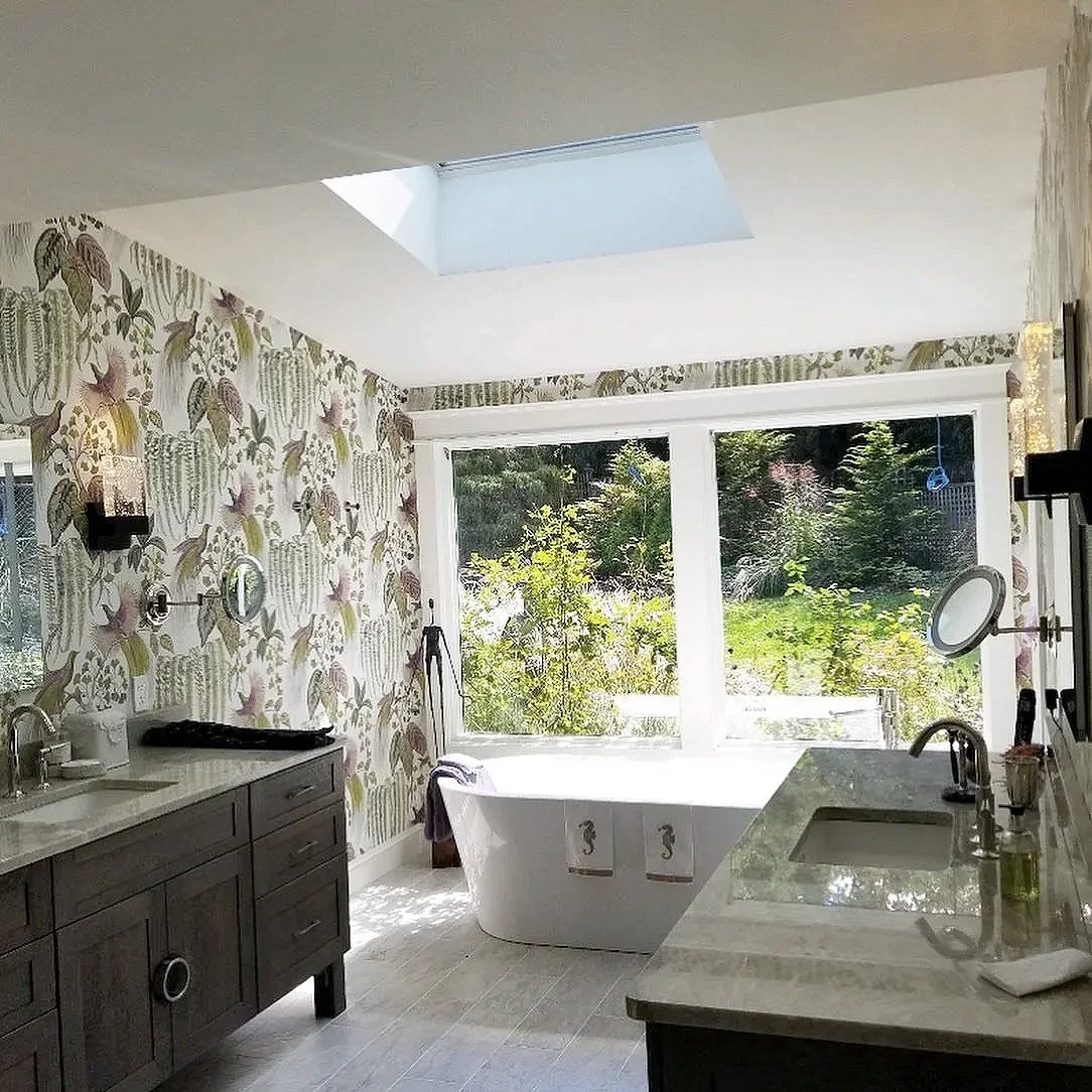 Bathroom with a large window showing greenery outside, a freestanding bathtub, a vanity with a sink, and a skylight in the ceiling.