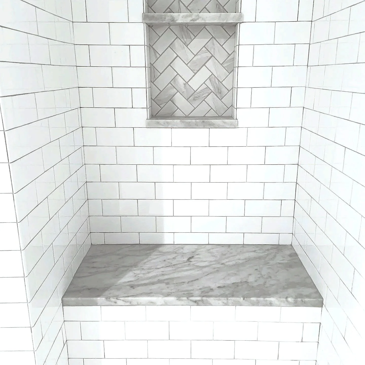 Shower niche with marble herringbone tiles and a marble shelf, surrounded by white subway tiles and a marble bench in a bathroom shower.