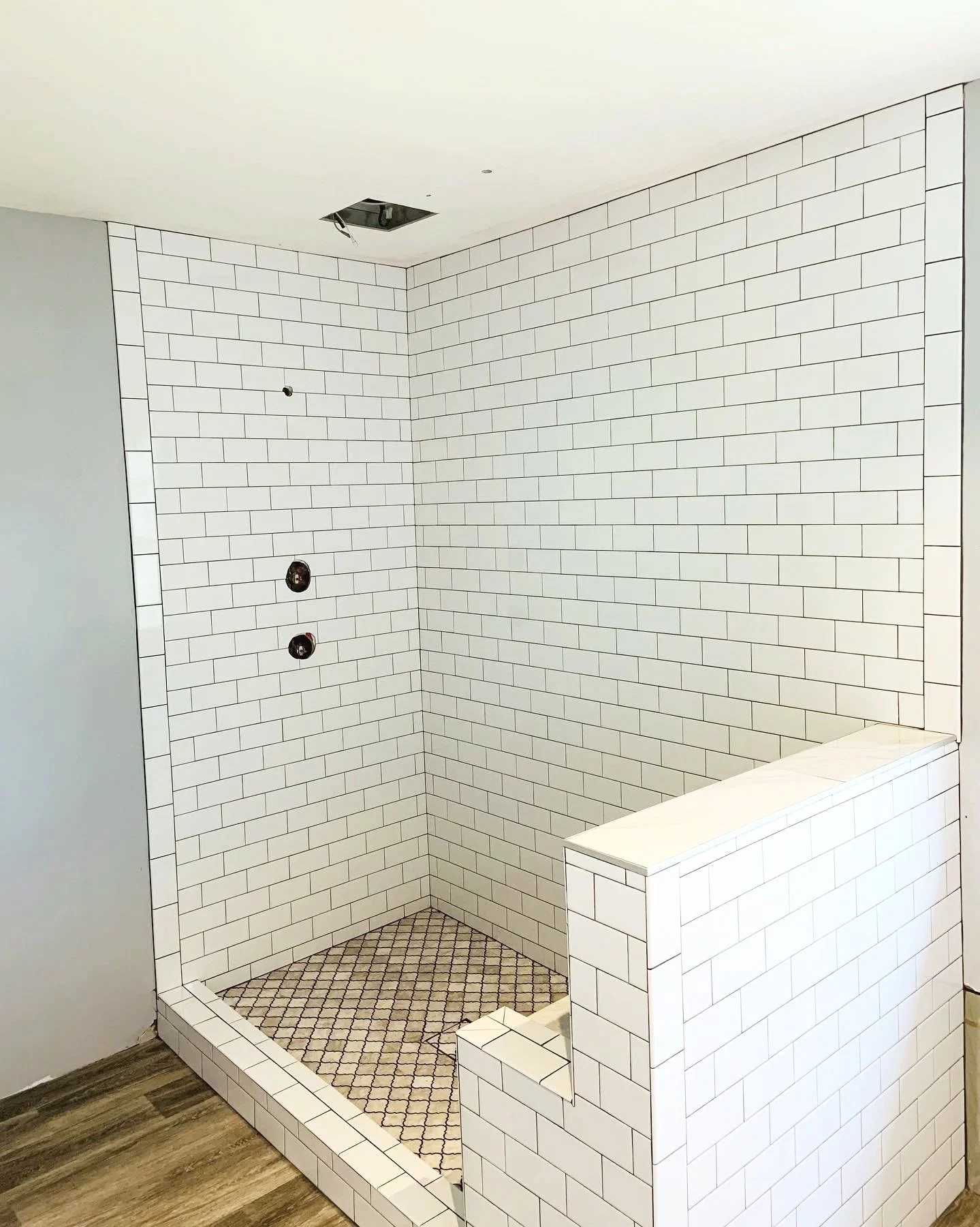 An unfinished shower with white subway tile walls, a small tiled bench, and a black hexagonal tile floor, with an open ceiling and exposed plumbing fixtures.
