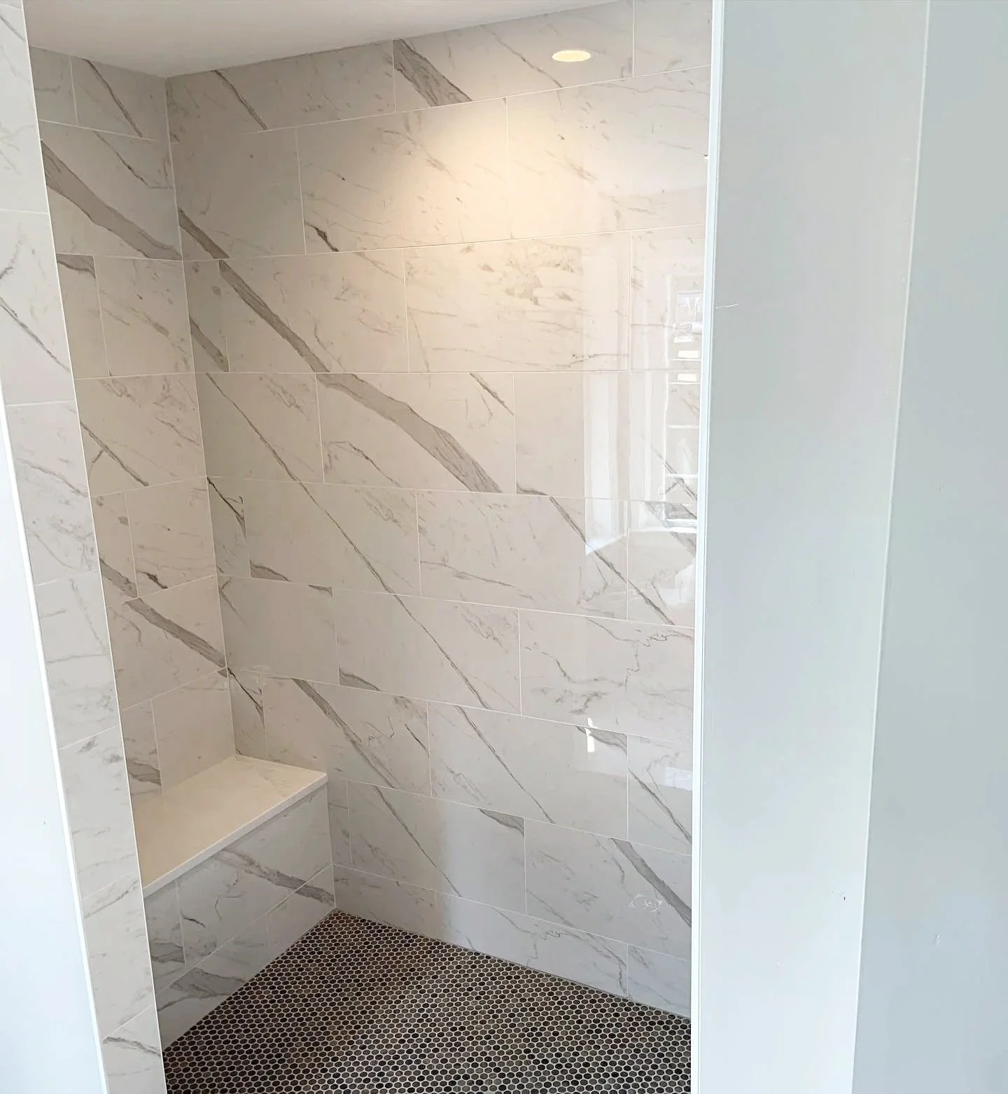 A modern shower with marble tile walls and a small built-in bench, featuring a dark hexagonal tile floor. There is a partial view of a window's reflection in the mirror.