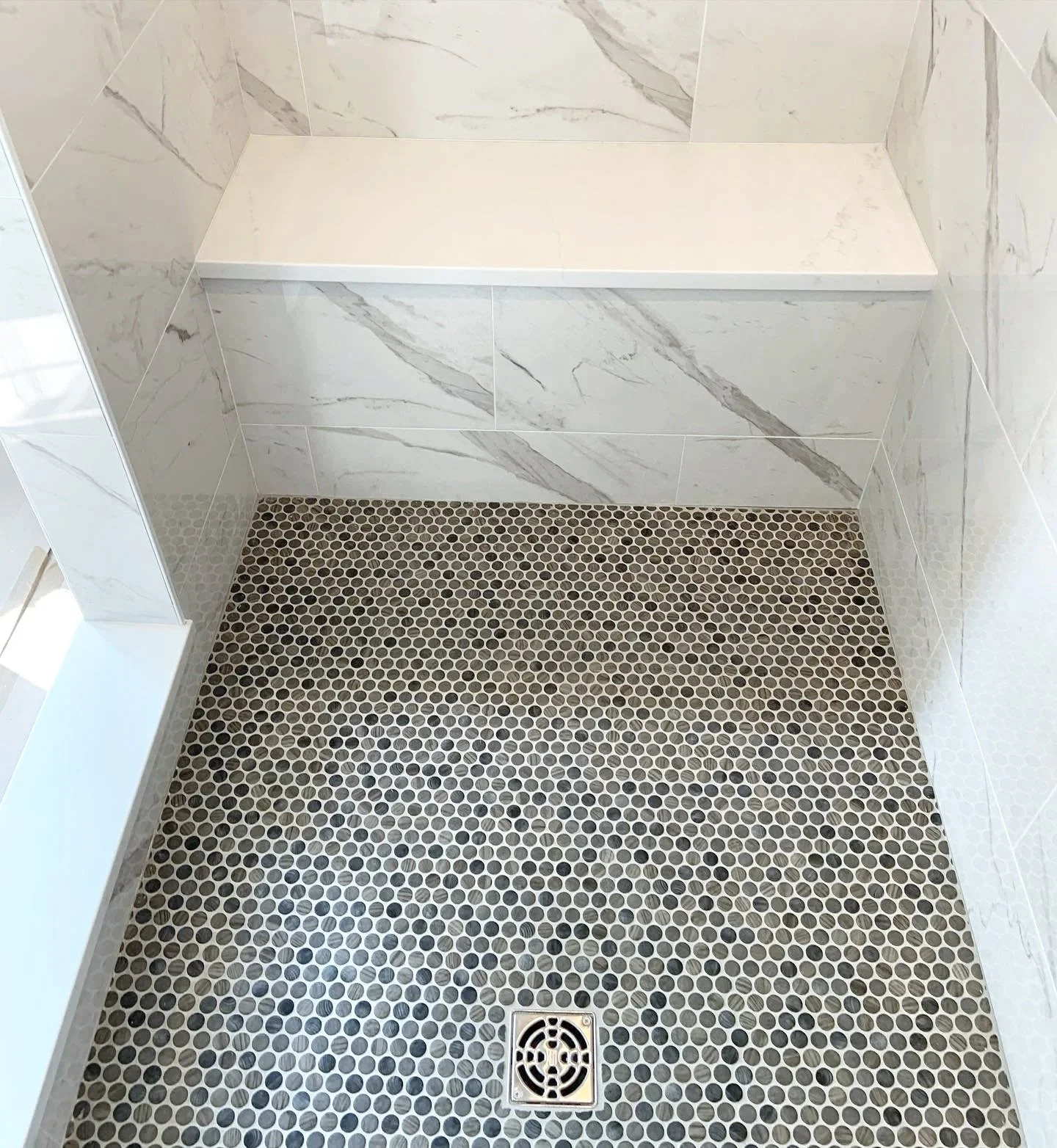 A walk-in shower with marble tile walls, a white shelf on the wall, a small round black pebble tile floor, and a square metal drain in the center.