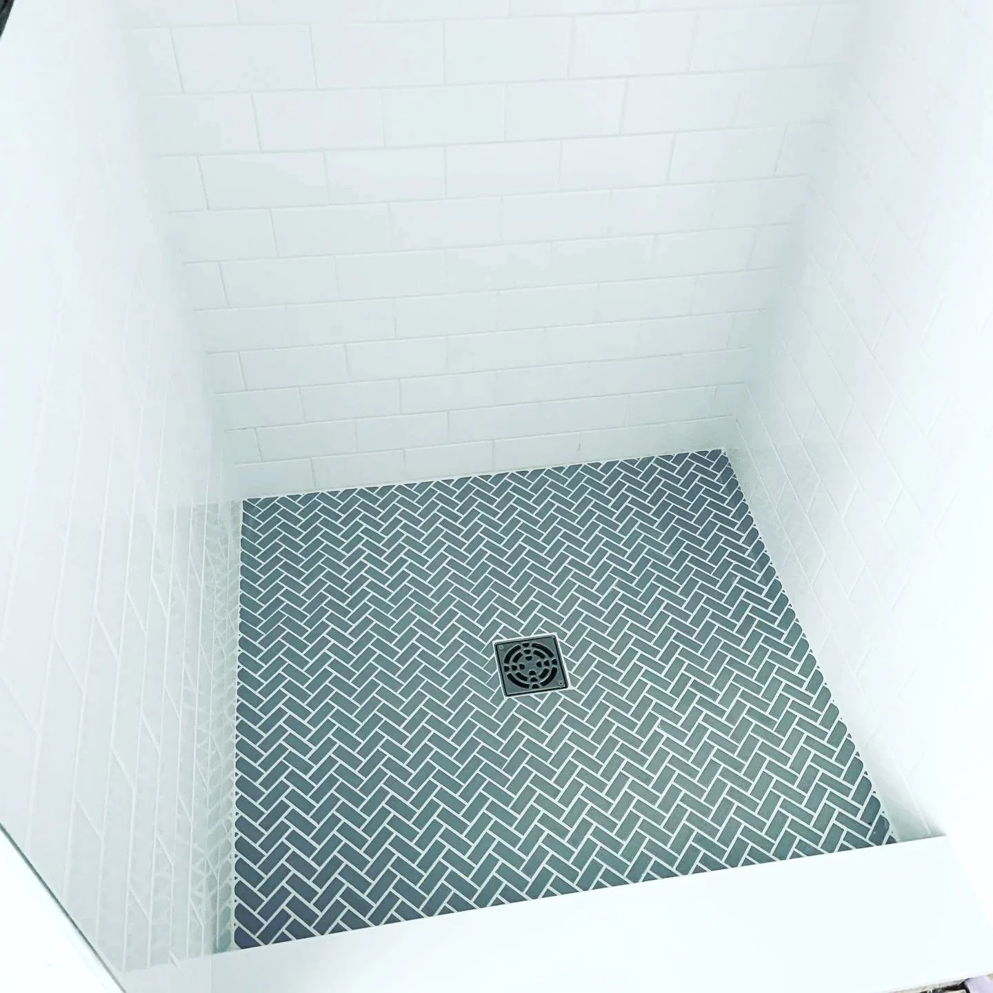 Empty shower stall with white subway-tile walls, dark herringbone-patterned floor tiles, and a central square drain.