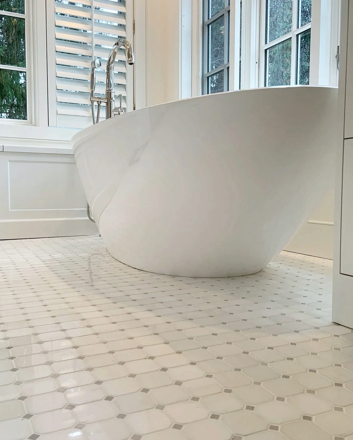 A white bathtub with a curved edge in a bathroom with large windows, white walls, and patterned tile flooring.