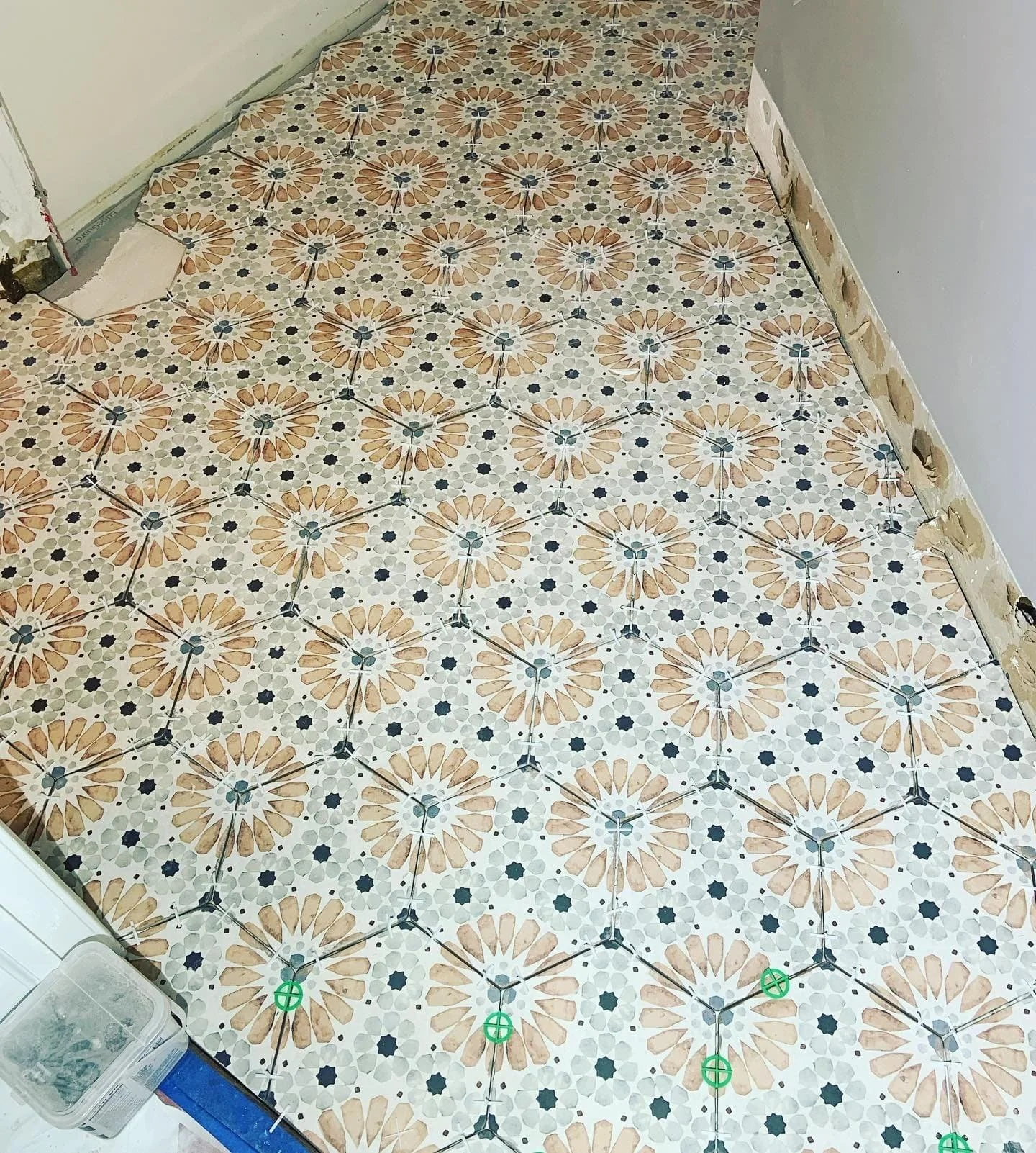 A close-up of a patterned tiled floor in a room under renovation. The tiles feature a floral design with tan, white, and black colors, and some areas of the floor are unfinished or damaged, especially near the wall on the right side.