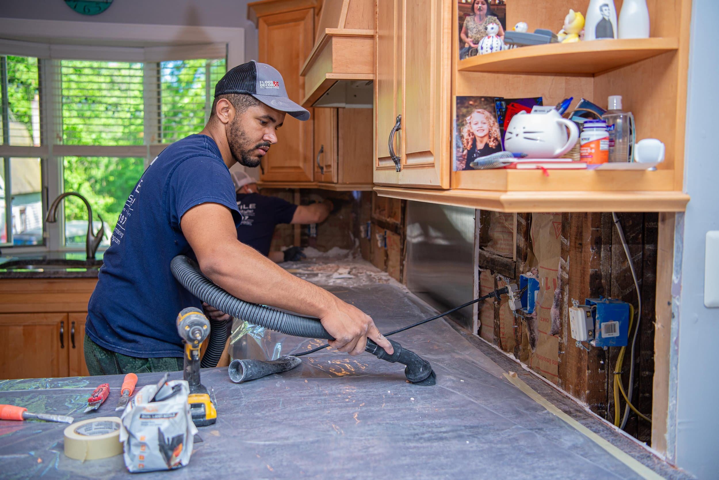 A man in a blue shirt and black trucker hat is working on installing or repairing kitchen cabinets, using a power tool on the counter. There are tools and materials on the counter, and the cabinet section is partially open, exposing wiring and insula