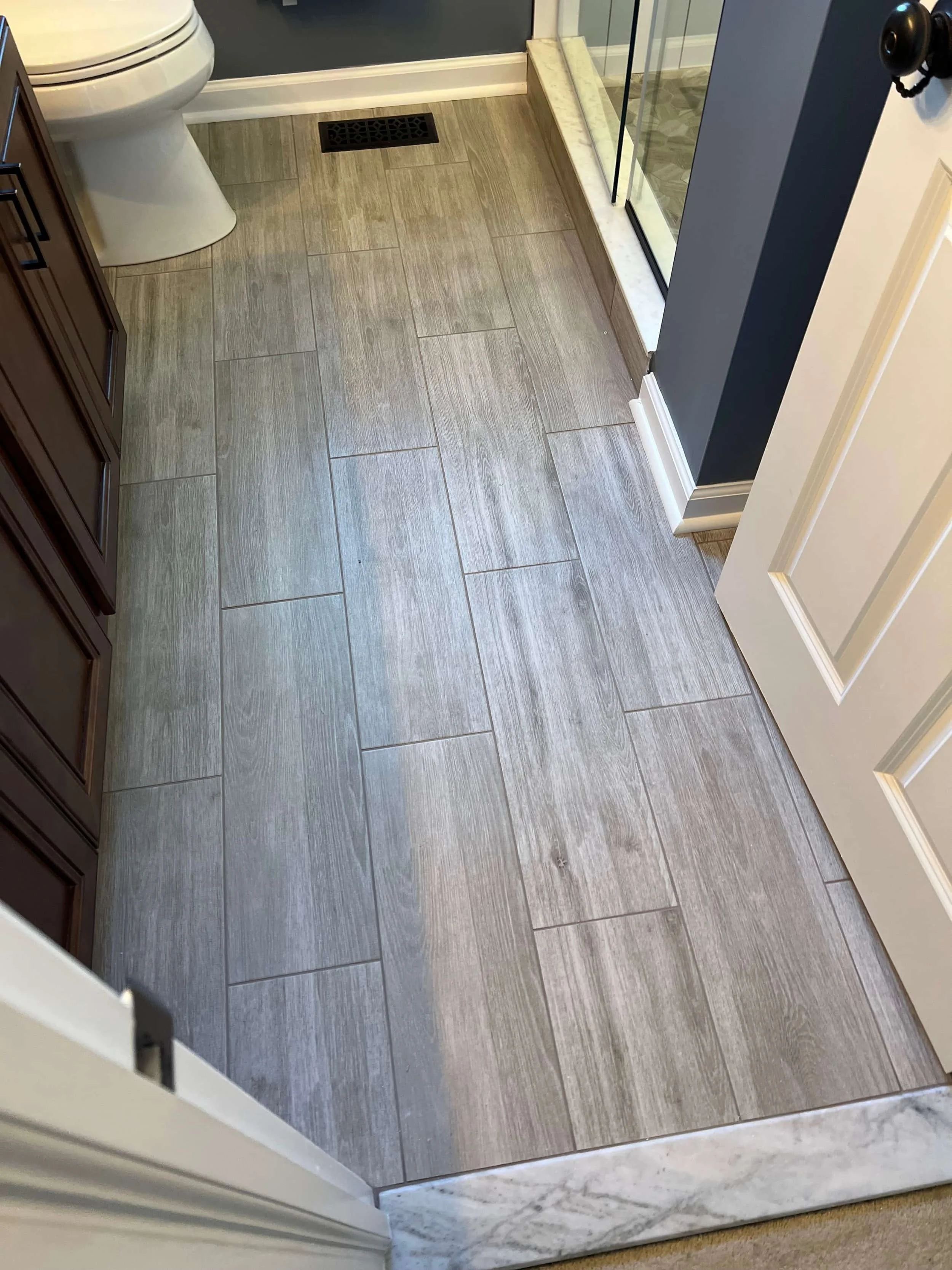 Floor of a bathroom with large gray tiles, adjacent to a marble threshold, a toilet in the corner, and a glass shower enclosure with a marble sill.
