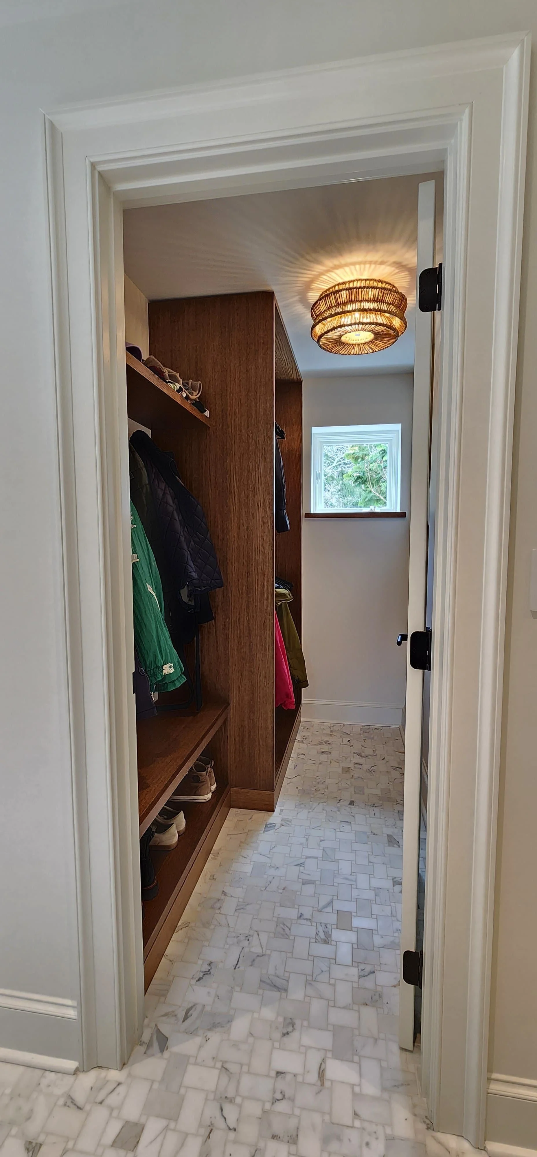 View of a small walk-in closet with a wooden shelving unit on the left, shoes on the bottom shelf, jackets and clothes hanging, and a small window at the end. The floor is covered with white and gray marble tiles, and a ceiling light fixture provides