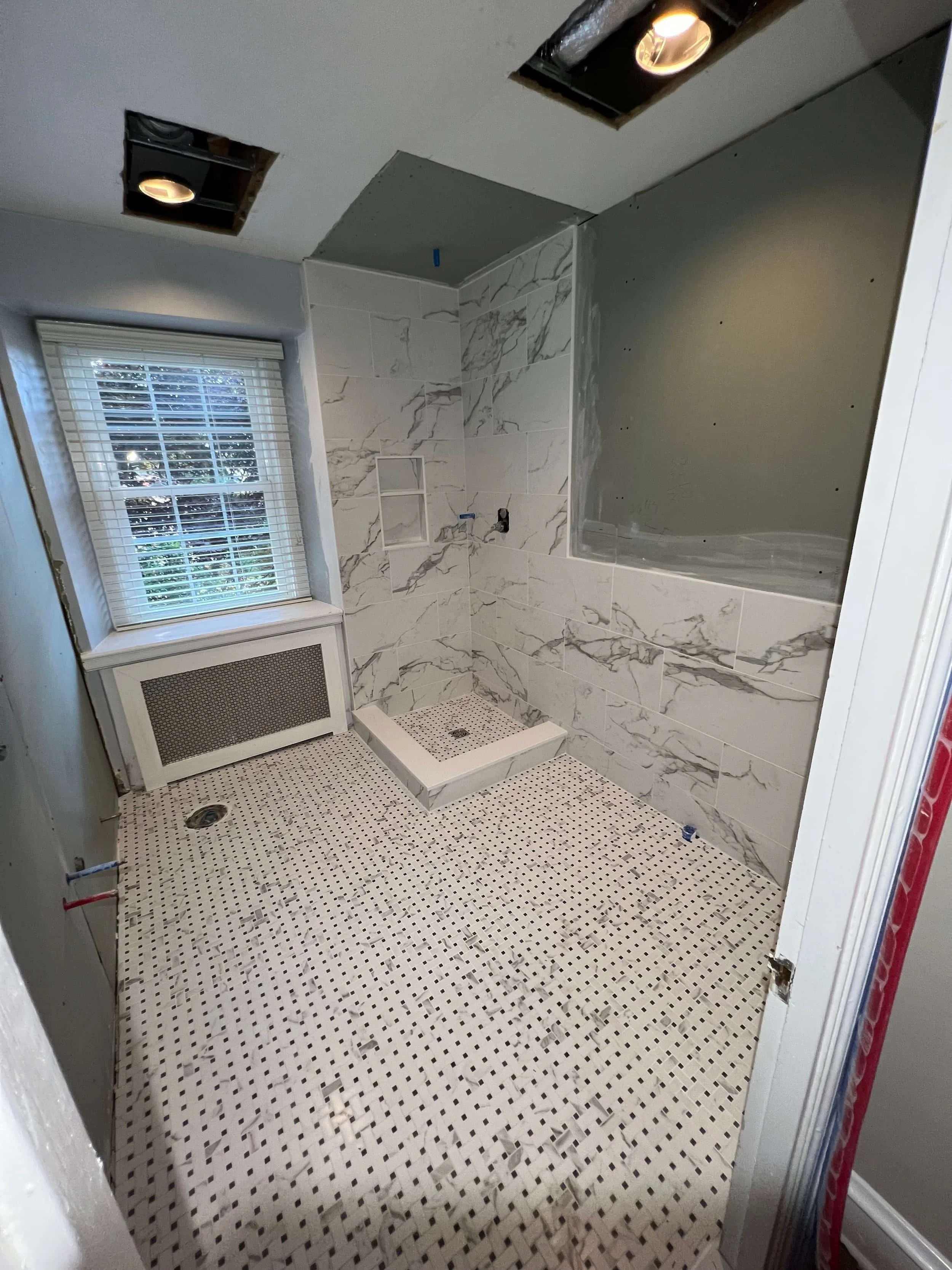 Under renovation bathroom with partially installed marble tile on walls, white and black mosaic tile on the floor, recessed lighting in ceiling, window with blinds, and a shower area under construction.