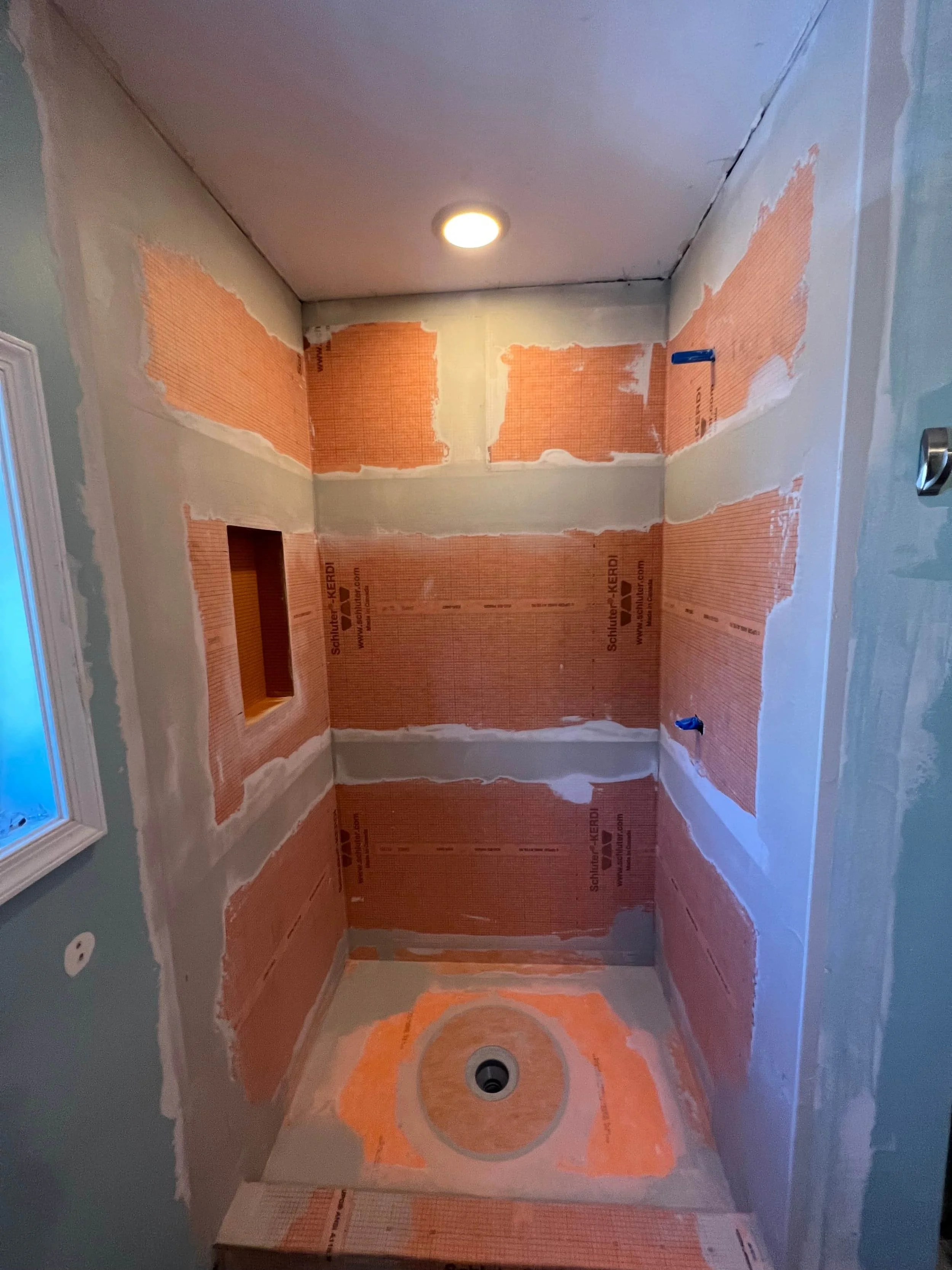 A bathroom with partially installed wall insulation and drywall, visible electrical outlets, a ceiling light, and an unfinished floor with a circular pattern, possibly for plumbing or drain installation.