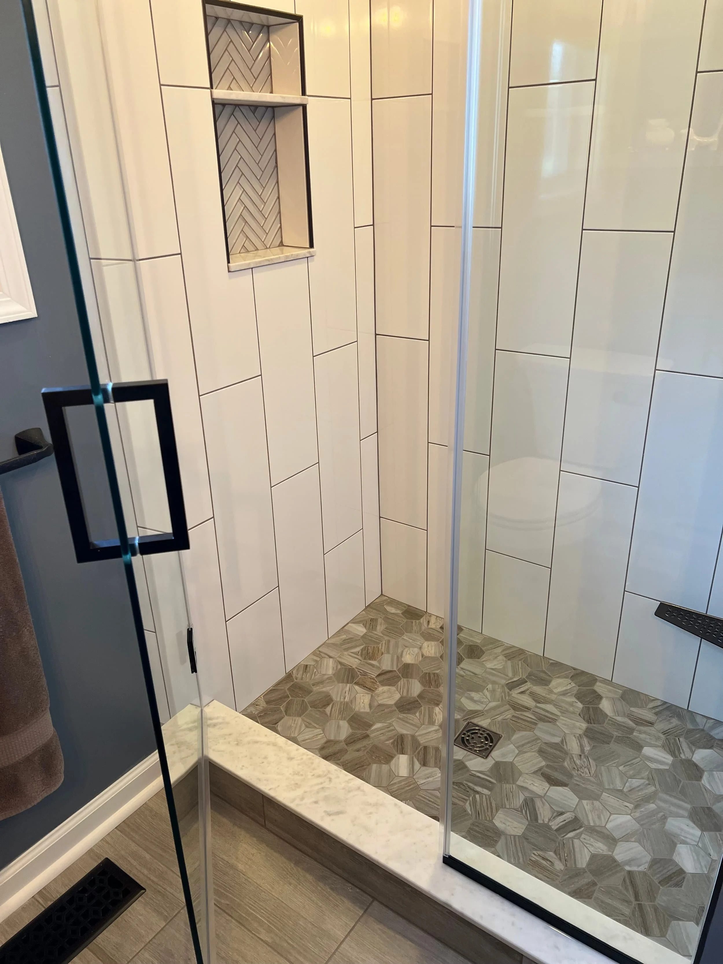 View of a modern bathroom shower with beige and gray tile walls, a built-in tiled shower niche, and a patterned tile floor. The shower has a glass door with a black handle and a black drain on the floor.