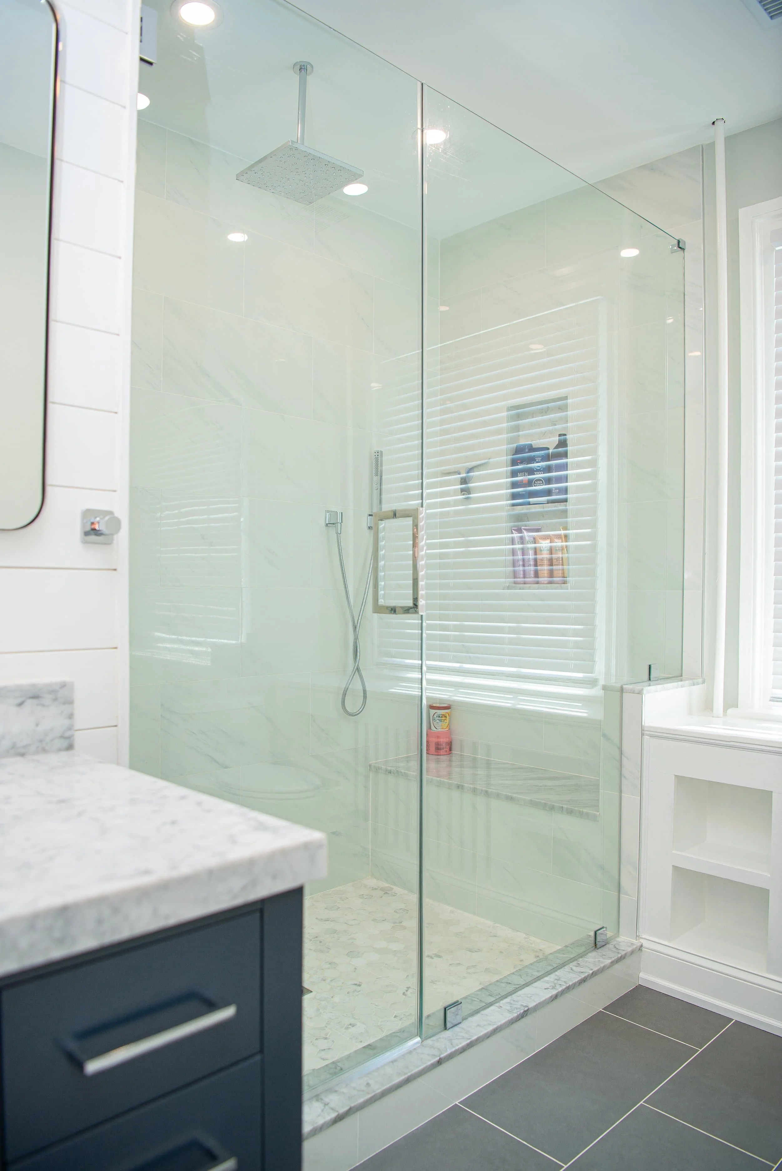 Modern bathroom with a glass-enclosed shower featuring a rainfall showerhead, white marble tiles, a built-in niche with toiletries, and dark gray floor tiles.