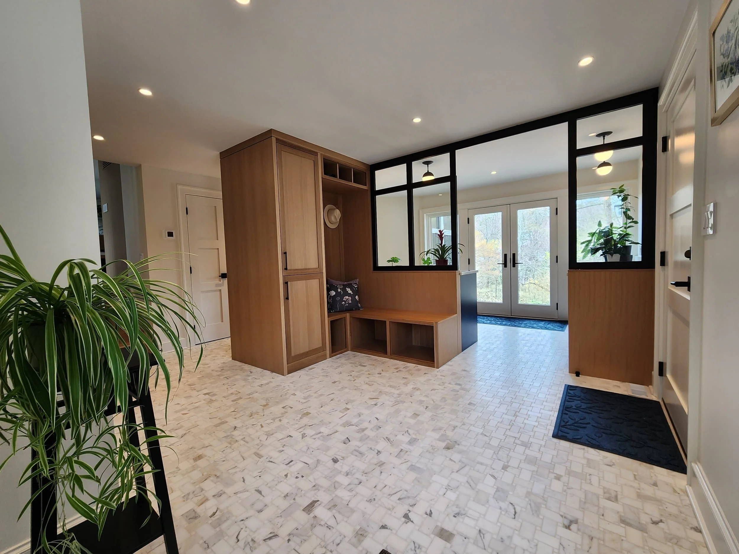 Interior entryway of a house with white and wood accents, black-framed glass doors, potted plants, and a seating nook with a hat on a built-in wooden bench.