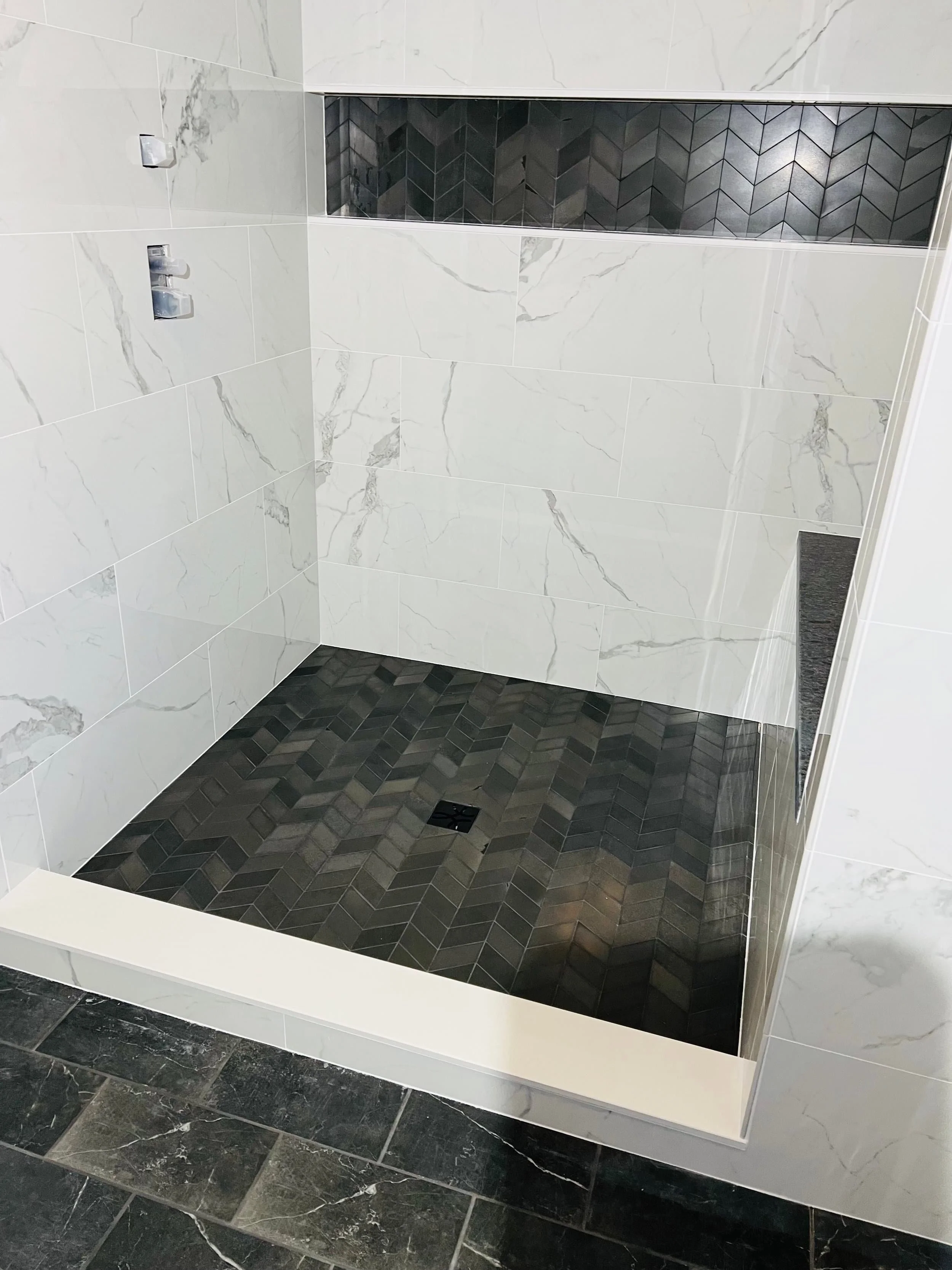 A modern shower with white marble wall tiles featuring grey veining, a built-in niche with black chevron tiles, and a black marble floor with a drain.