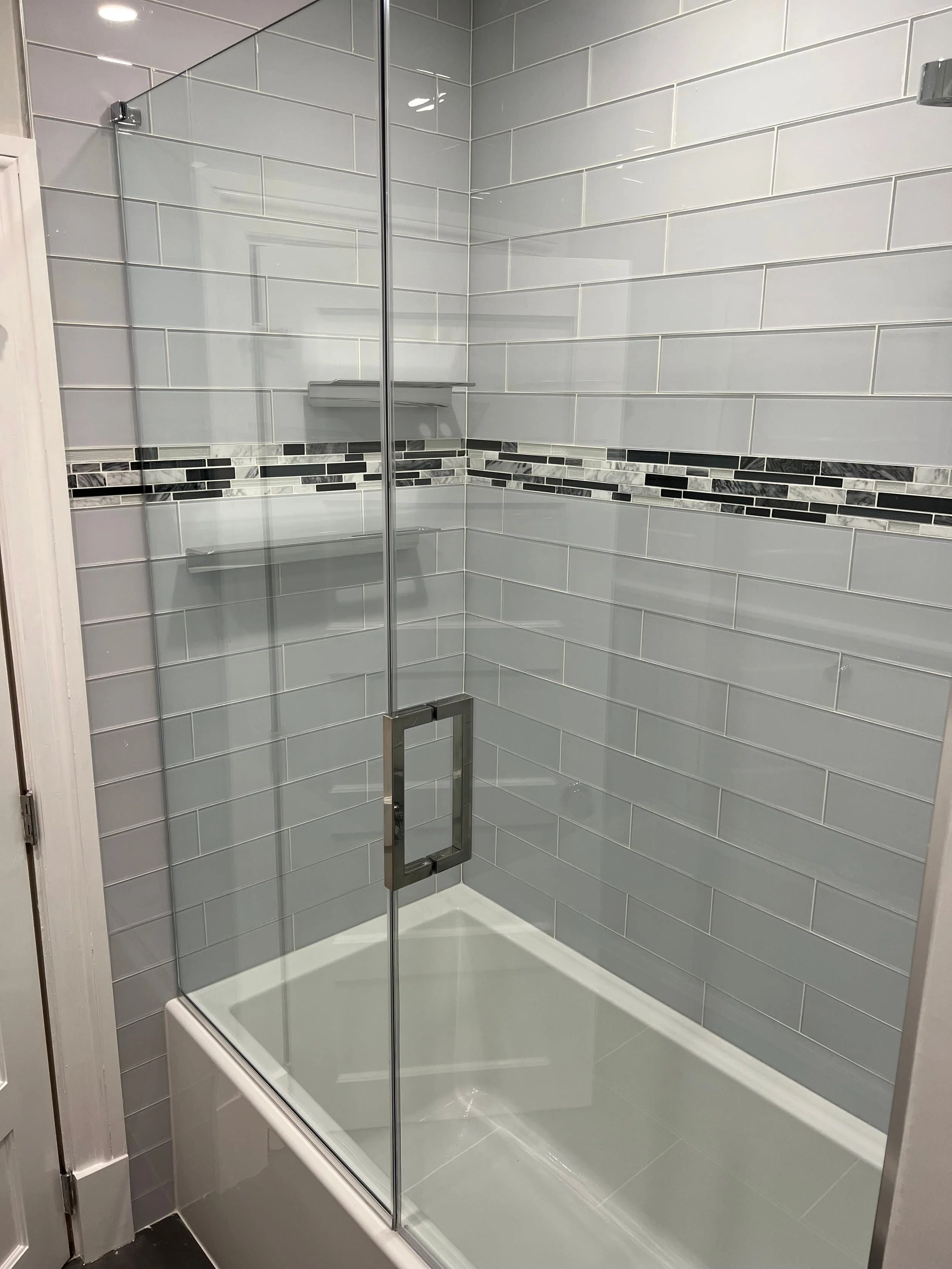 A modern shower with glass enclosure, gray subway tiles, and a decorative strip of black and white mosaic tiles.