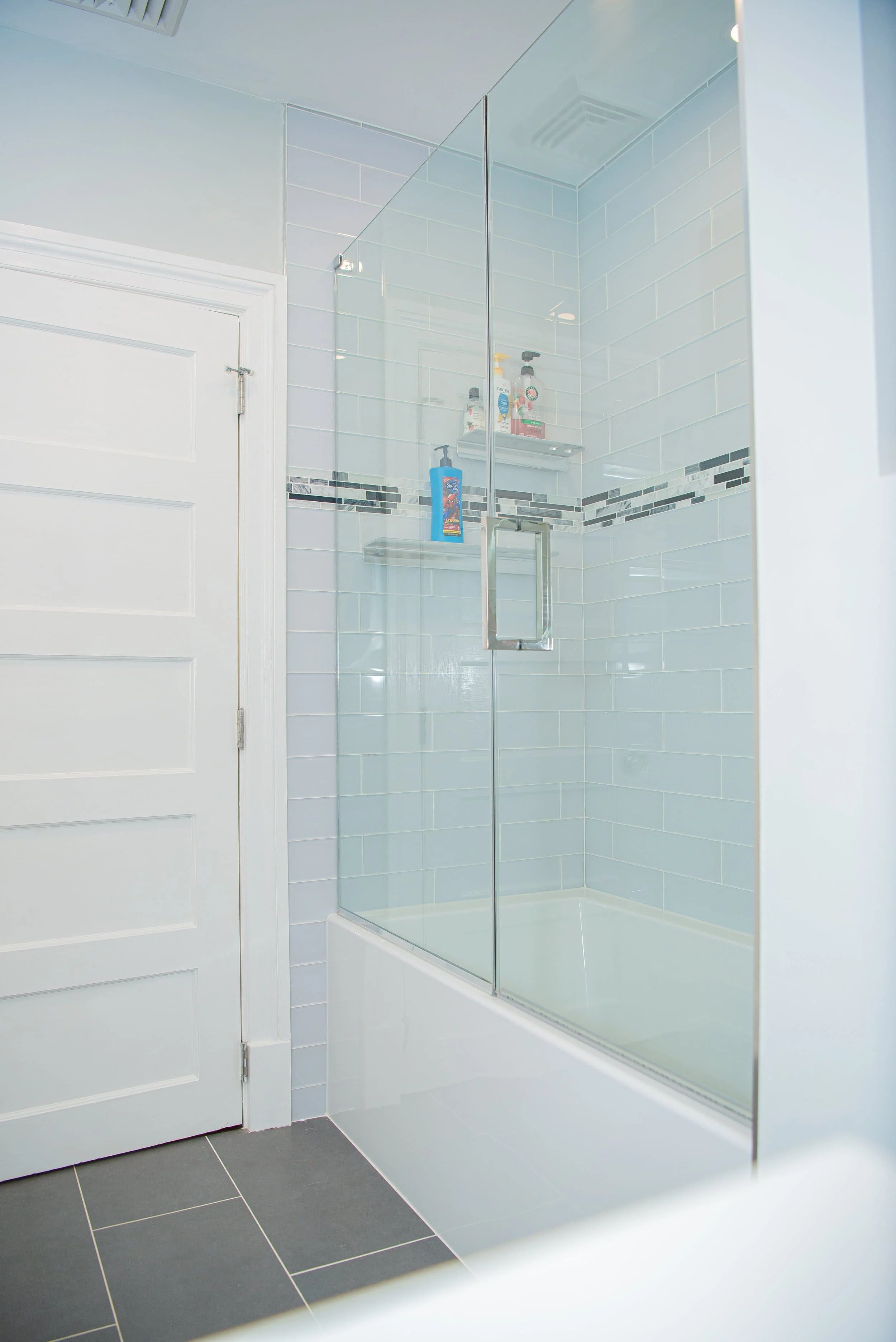 A bathroom shower area with clear glass enclosure, white tiles with gray and black mosaic border, and toiletries on a shelf. The floor has dark gray tiles, and there is a white door nearby.