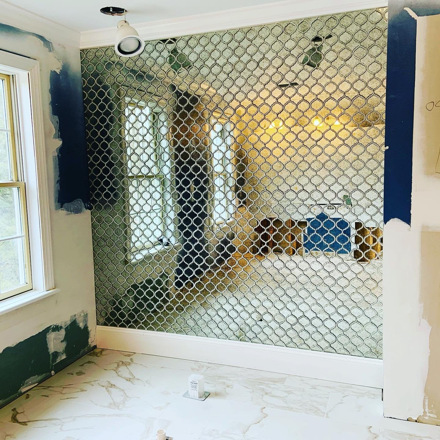 A room undergoing renovation with a partial wall painted dark blue, a window, white marble-like tile flooring, and a decorative glass partition with a geometric pattern.