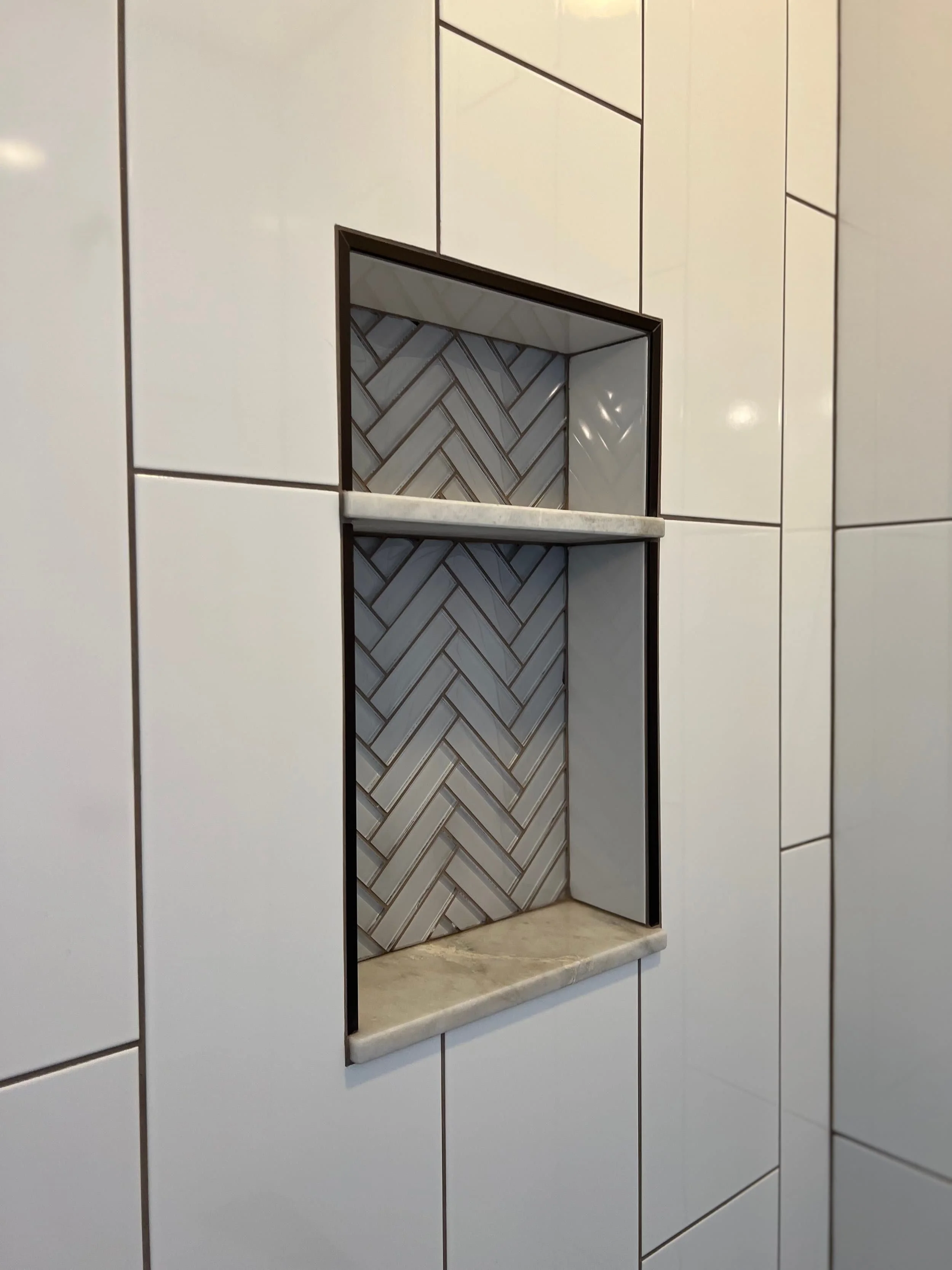 Close-up of a built-in wall niche with a white marble shelf and herringbone-patterned tile interior, surrounded by white wall tiles.