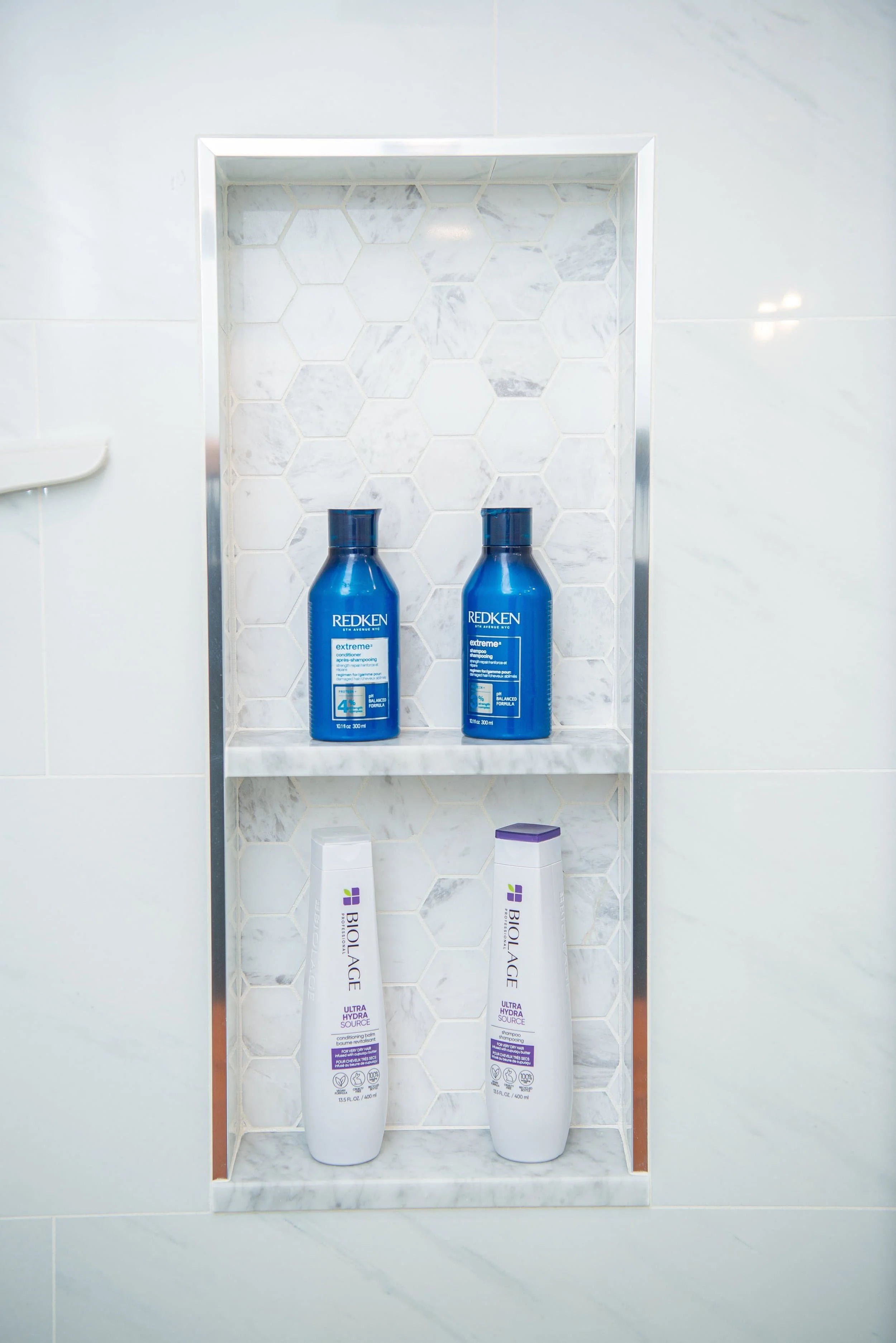 Shower niche with marble tiles containing two blue bottles of Redken shampoo and two white bottles of Biolage hair care products.