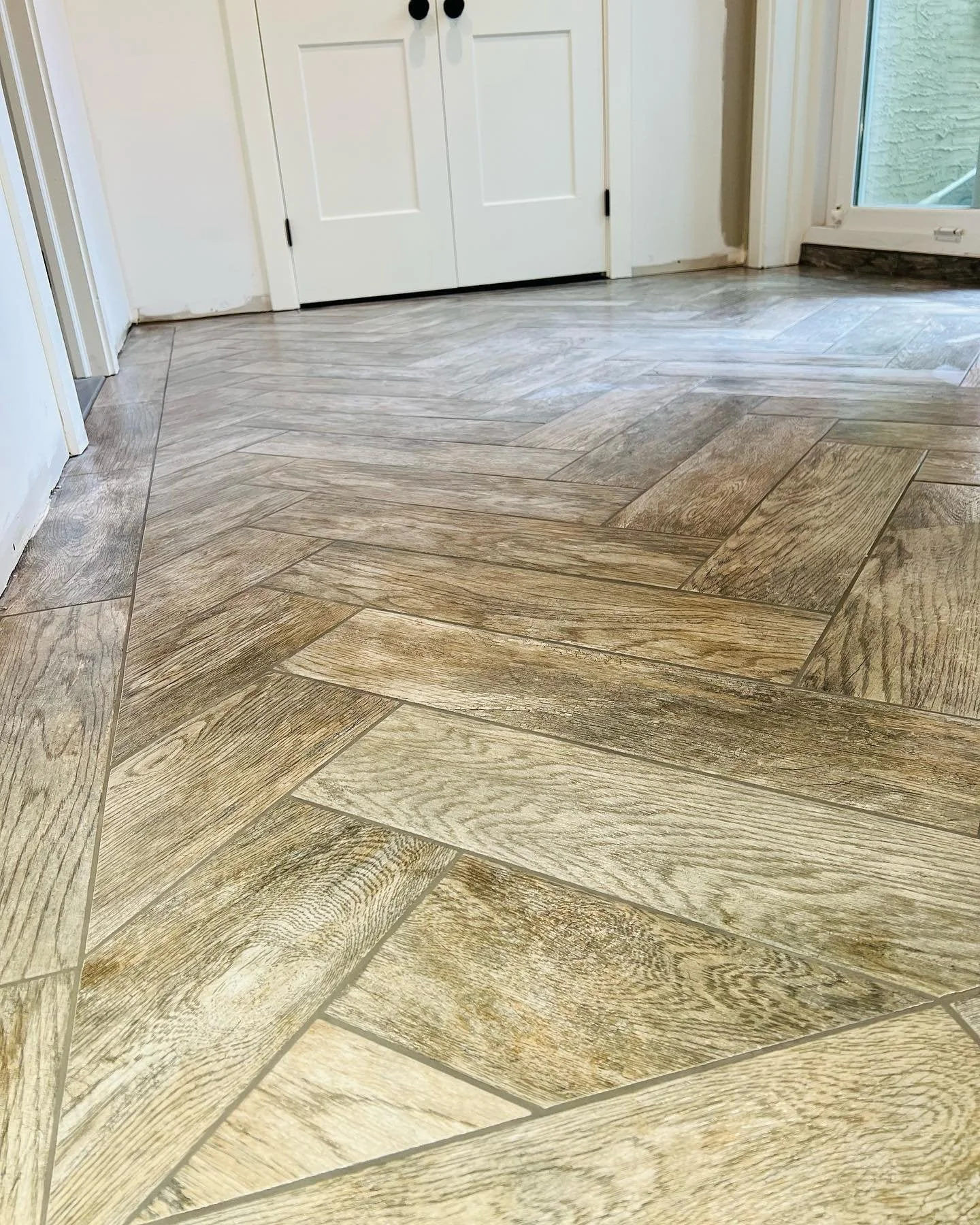 Wood-look tile flooring in a herringbone pattern inside a house near a white door with black doorknobs and a sliding glass door leading outside.