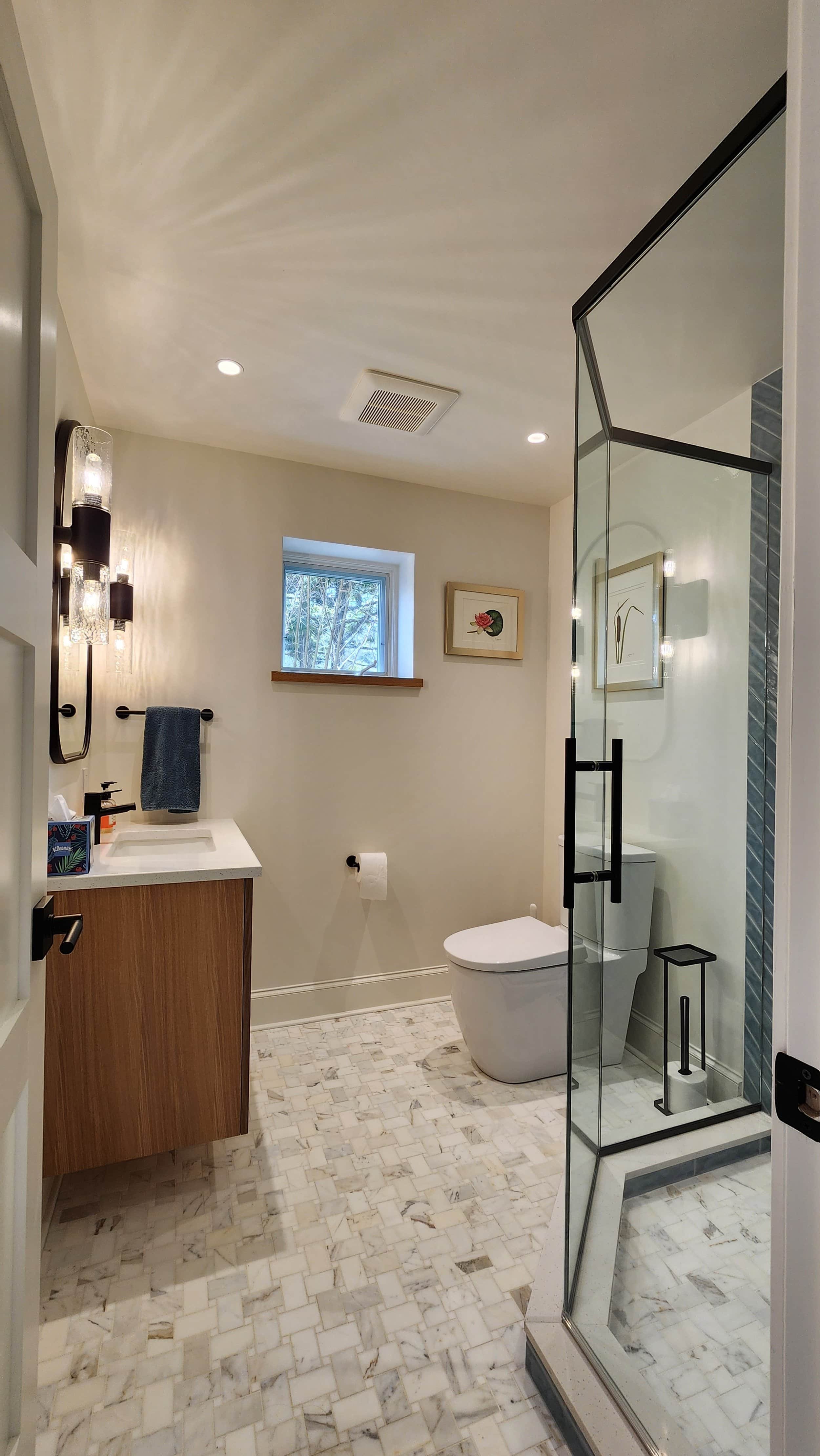 A modern bathroom with a small window, artwork on the wall, marble tile flooring, a glass shower enclosure, a toilet, a wooden vanity with a sink, and wall-mounted light fixtures.