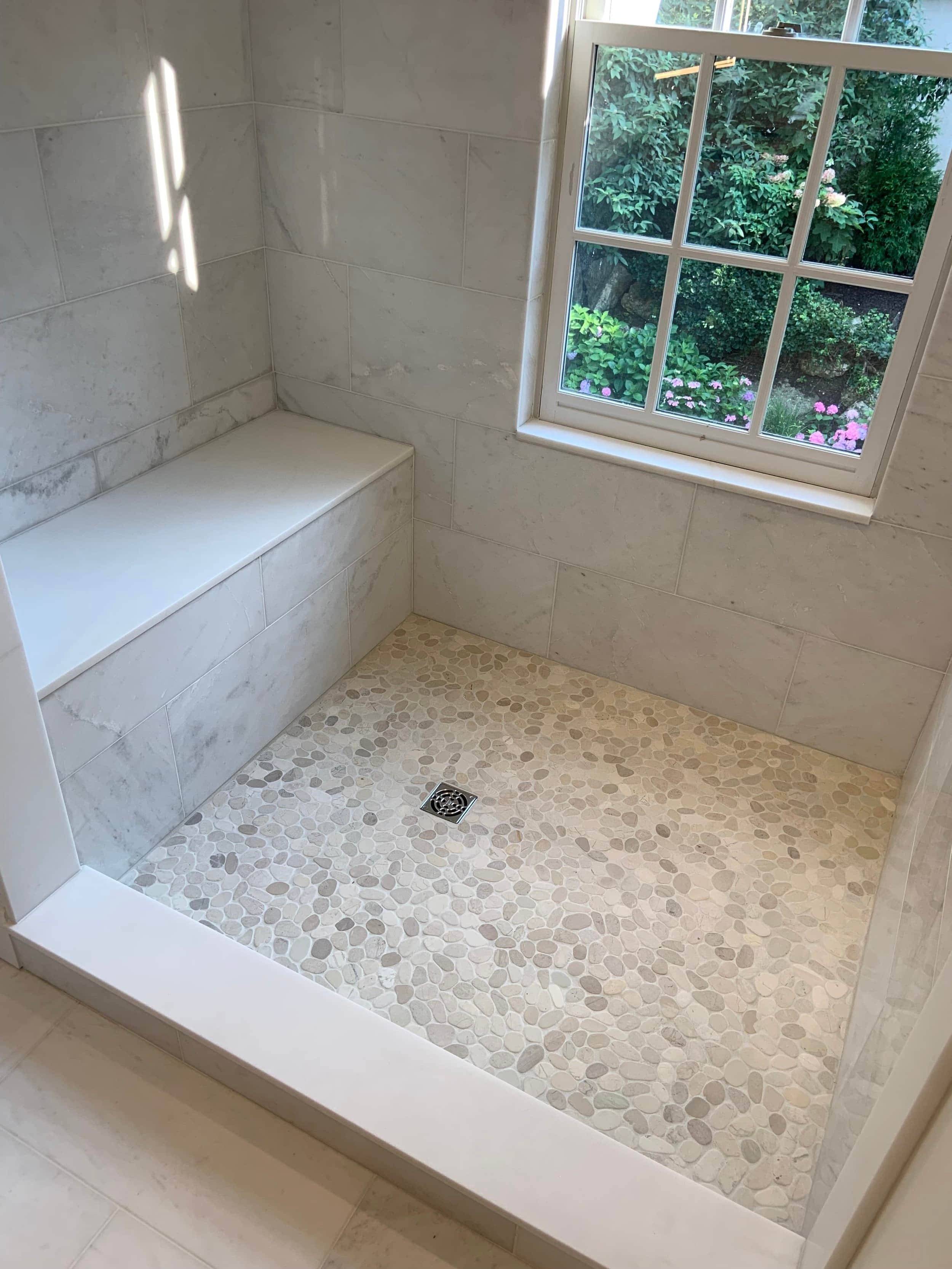 A shower area with beige pebble tile floor, light-colored wall tiles, a built-in bench, a drain on the floor, and a window showing greenery outside.