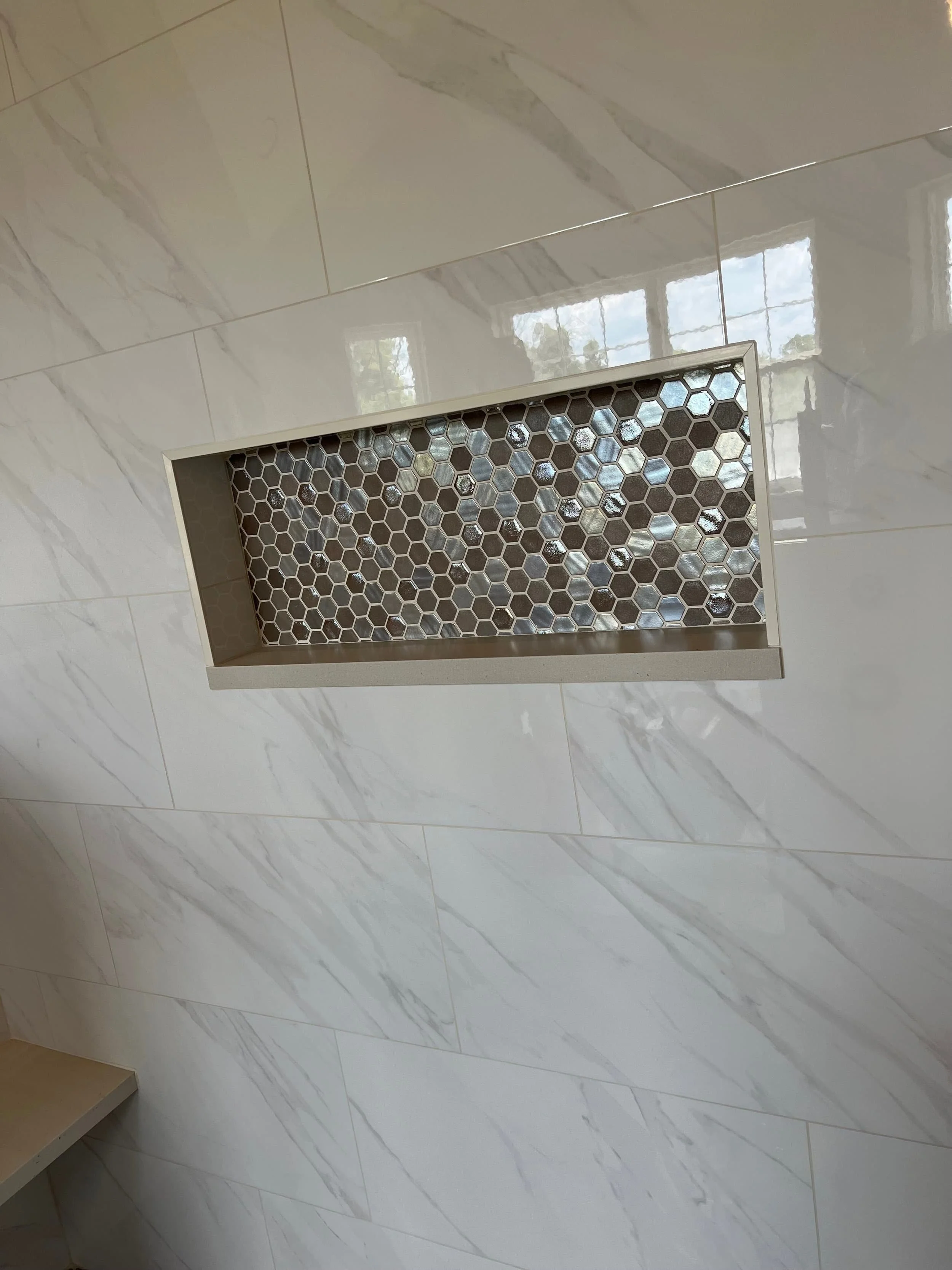 Close-up of a modern bathroom wall with large white marble tiles and a recessed decorative niche with small hexagonal tiles and glass stones inside.