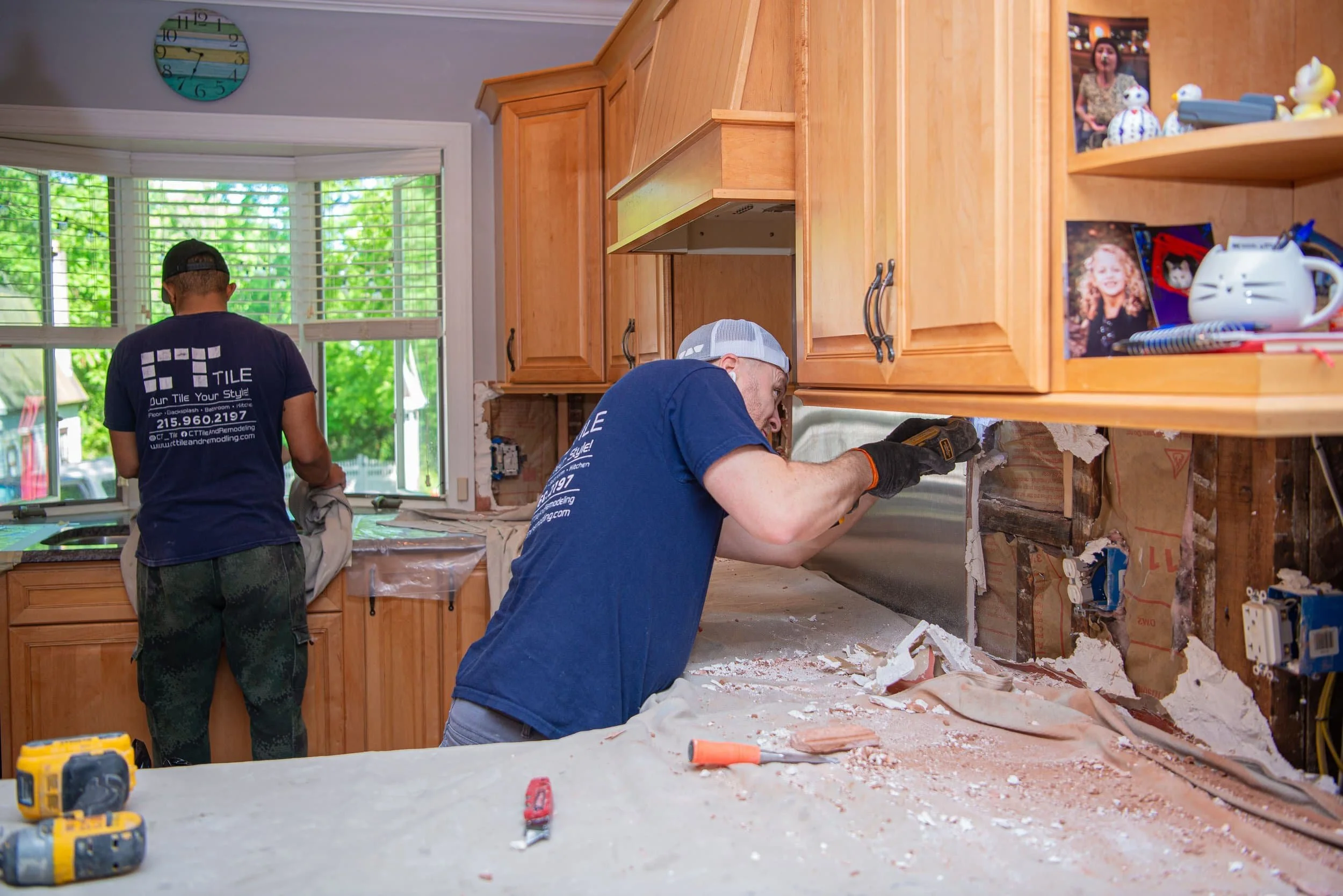 Two men working on kitchen renovation, one removing cabinetry and the other working underneath, with tools on a work surface and a window with blinds showing trees outside.