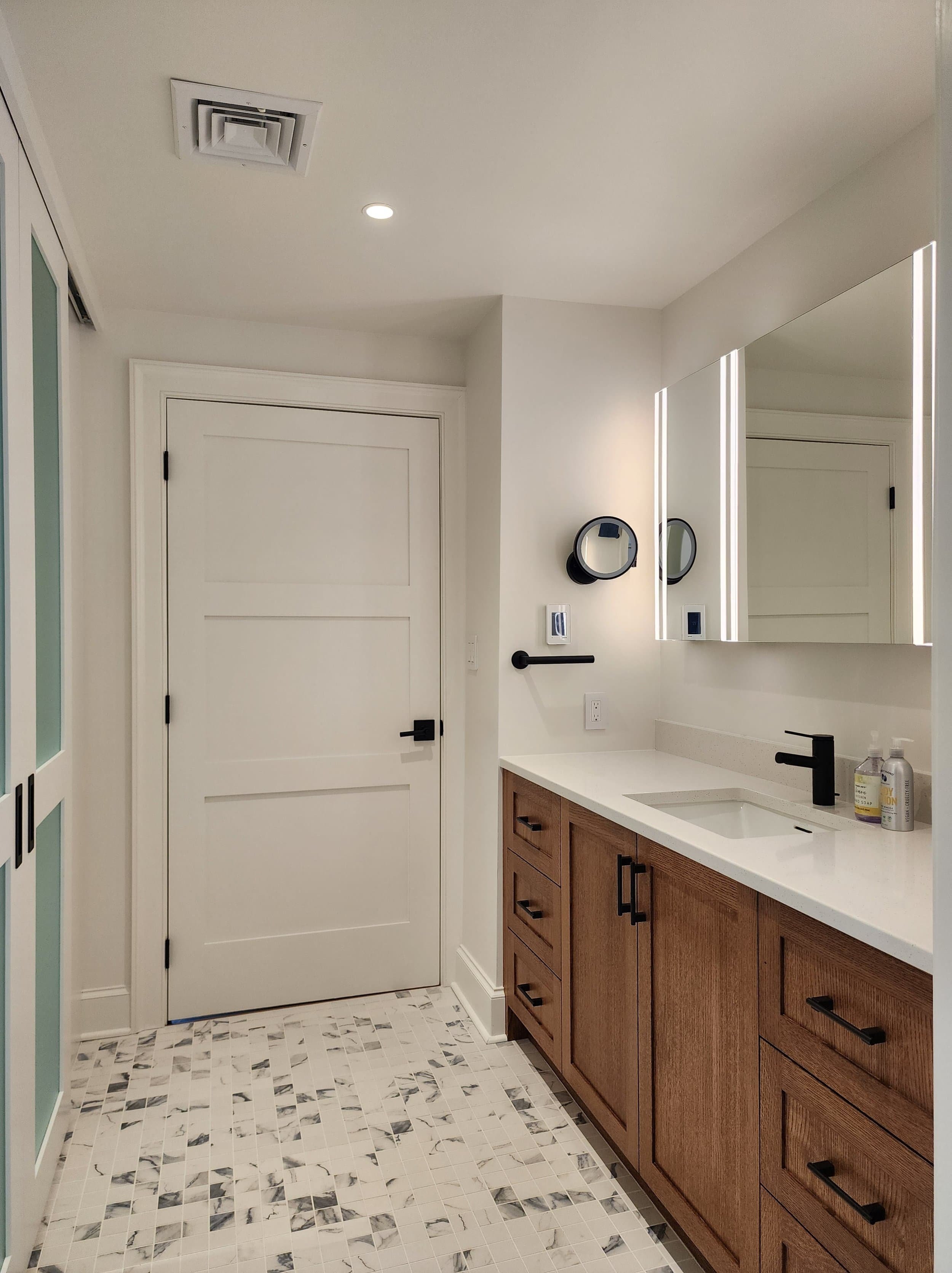 A modern bathroom with white walls and a white door. There is a wooden vanity with black hardware, a white countertop, and a black faucet. A large mirror with built-in LED lighting is mounted above the vanity. The floor has a geometric patterned tile