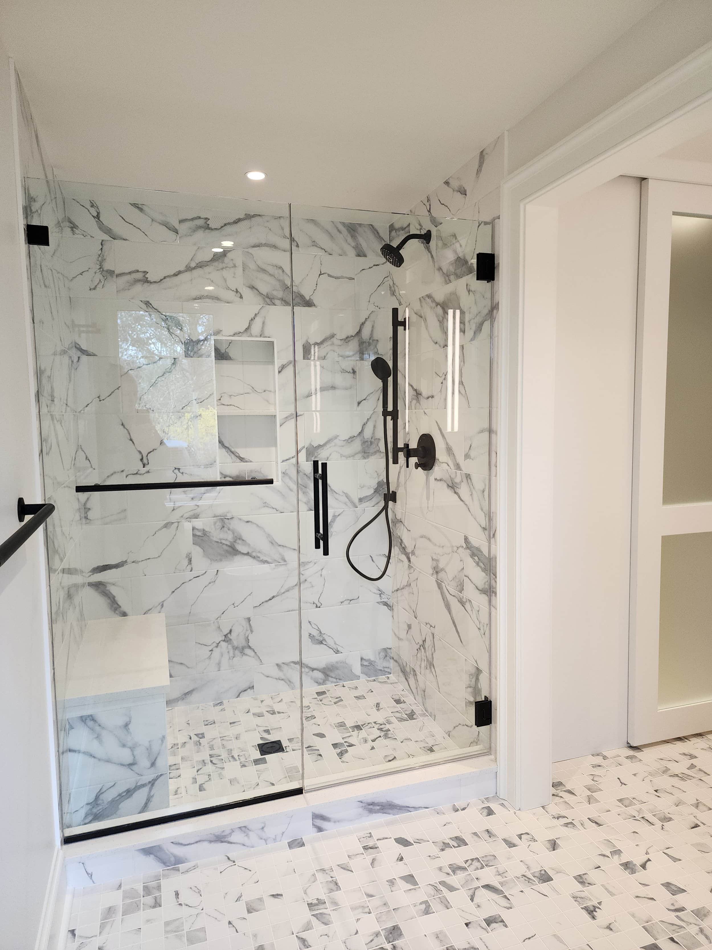 Modern walk-in shower with white marble tiles and black fixtures, including a rain showerhead and a handheld shower. The shower has glass doors, and the floor features small square marble tiles, with a drain at the center.